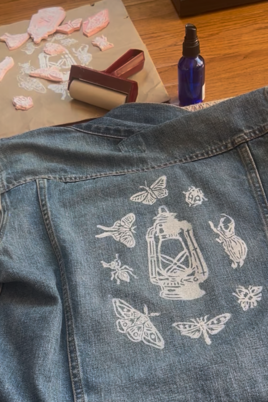 Back of a denim jacket with white butterfly and lantern design, on a wooden surface with pink soaps, a roller, and a spray bottle nearby.