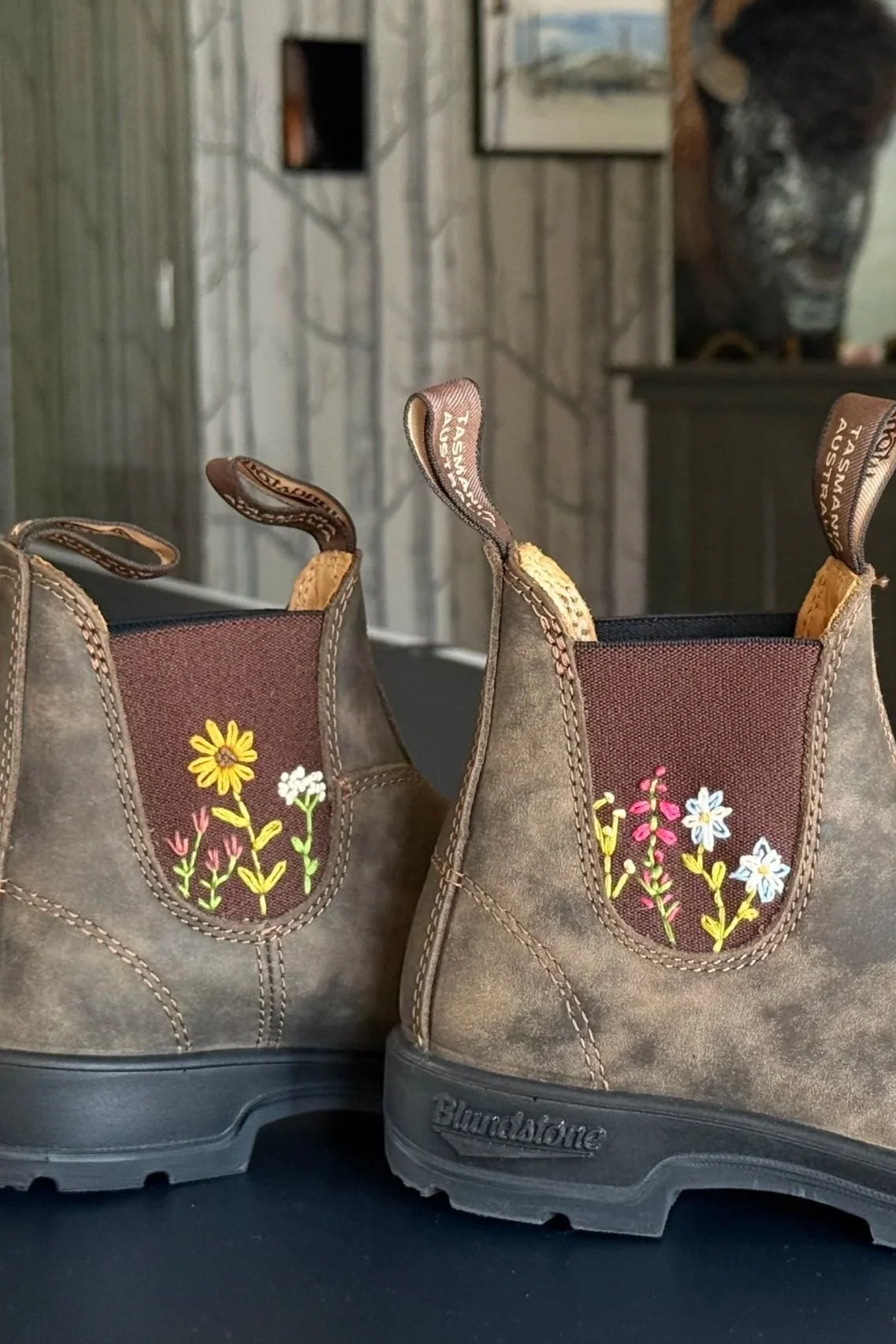 Pair of boots with embroidered colorful flowers on the sides, set on a black surface with a rustic wood-paneled background.