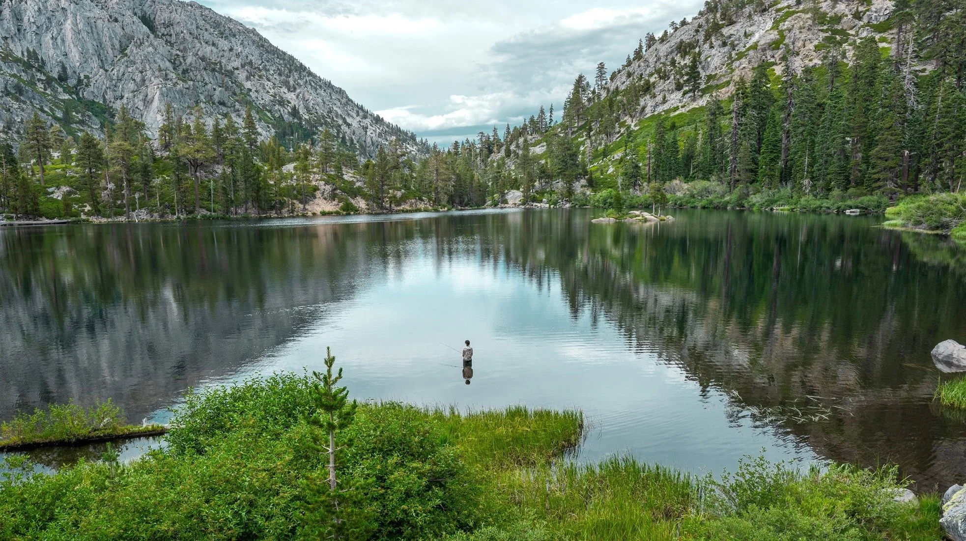 A person fishing in a calm mountain lake surrounded by green trees and rocky cliffs under a cloudy sky.