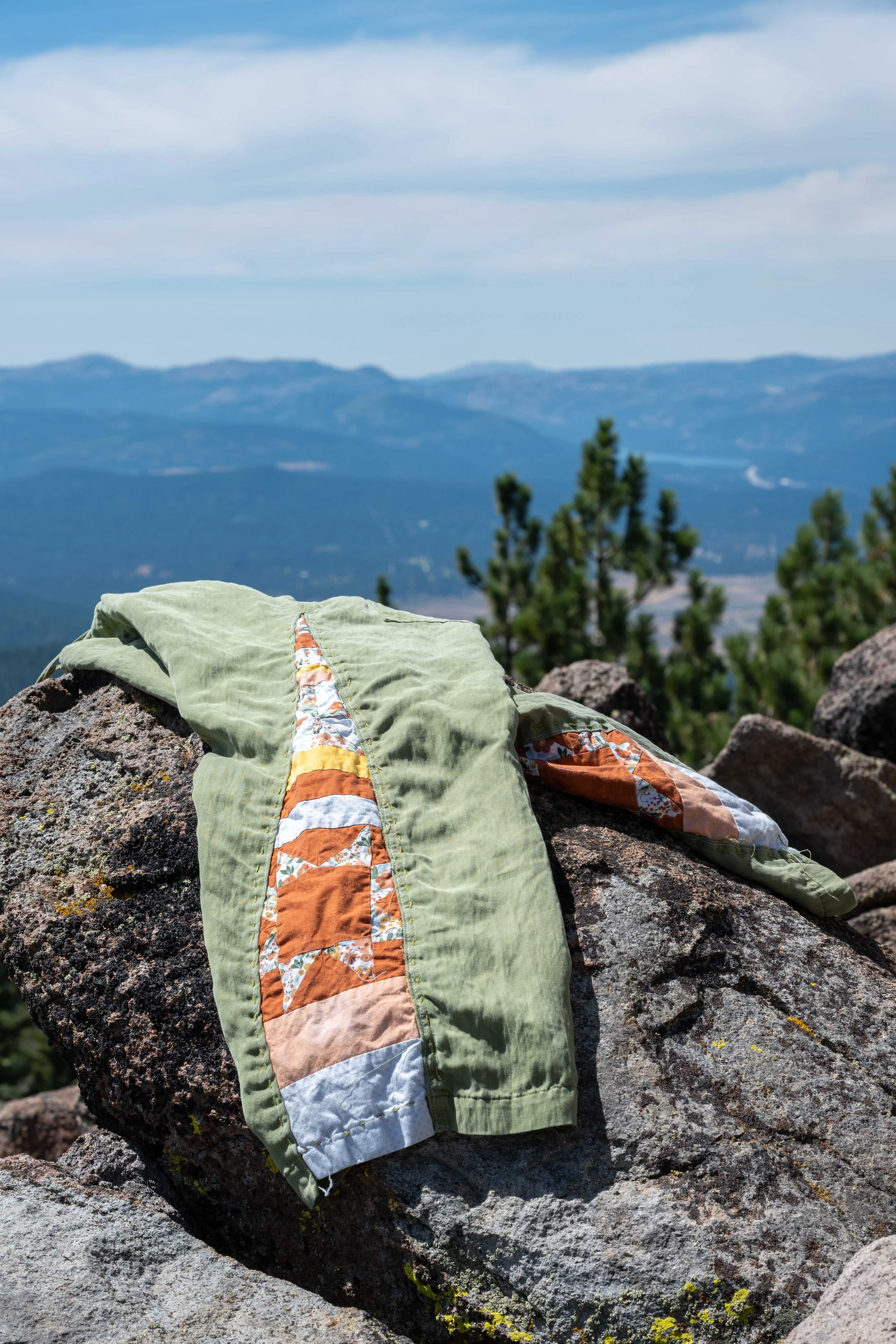 A pair of hiking pants with a colorful quilted stripe running down the side, draped over a large rock in a mountainous landscape with pine trees and distant mountains under a partly cloudy sky.