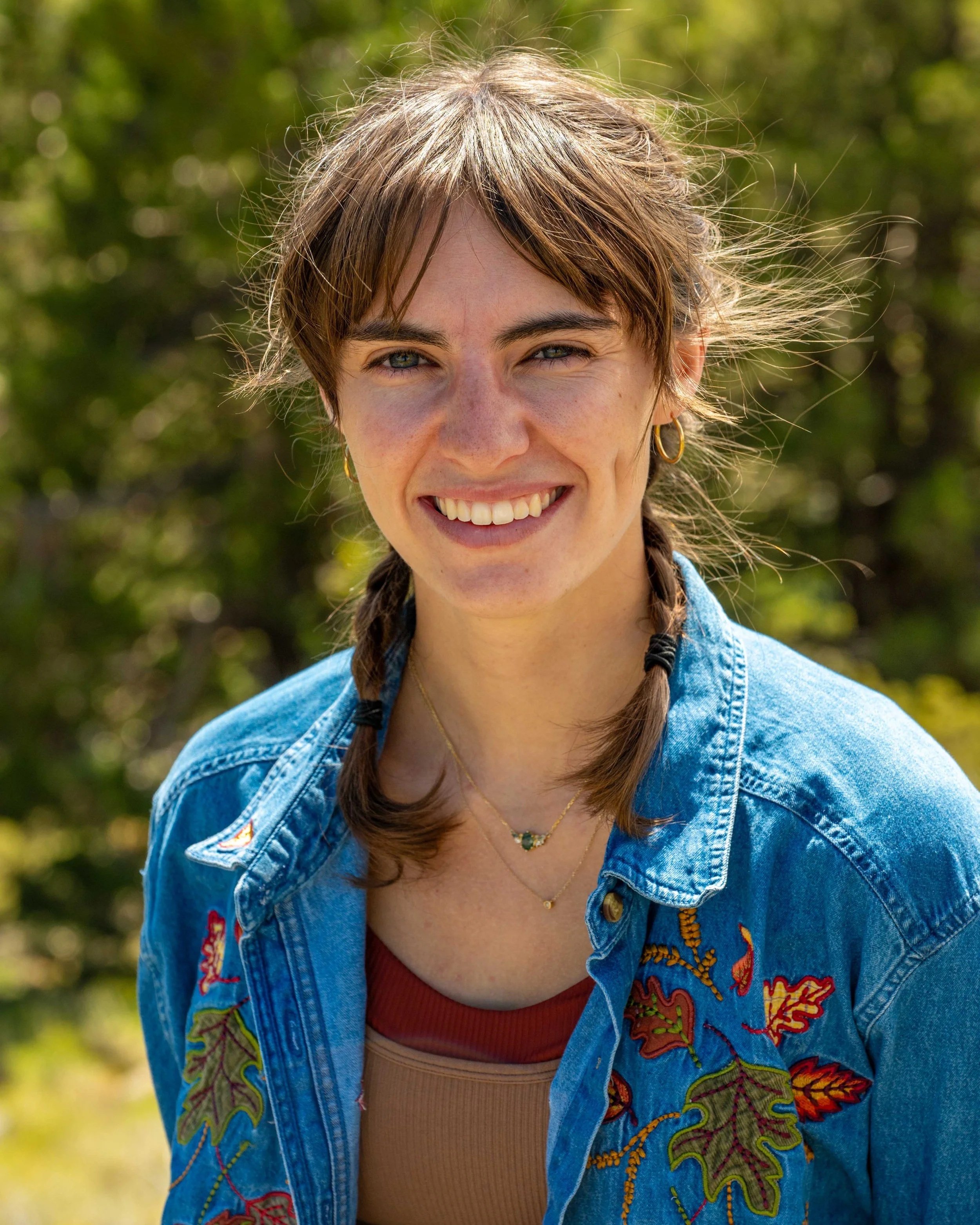 A smiling woman with brown hair, wearing a blue embroidered denim jacket, a gold necklace, and gold earrings, outdoors with a blurred green background.