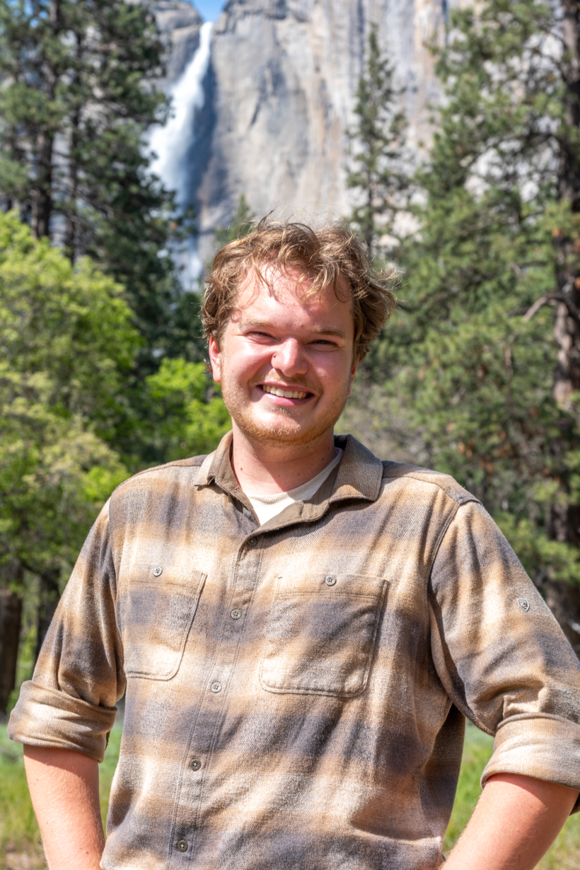 A young man with light brown curly hair and a beard smiling outdoors with a waterfall and green trees in the background.