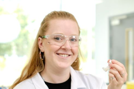 A woman with red hair, glasses, and earrings smiling in a lab or medical setting, holding a small object.