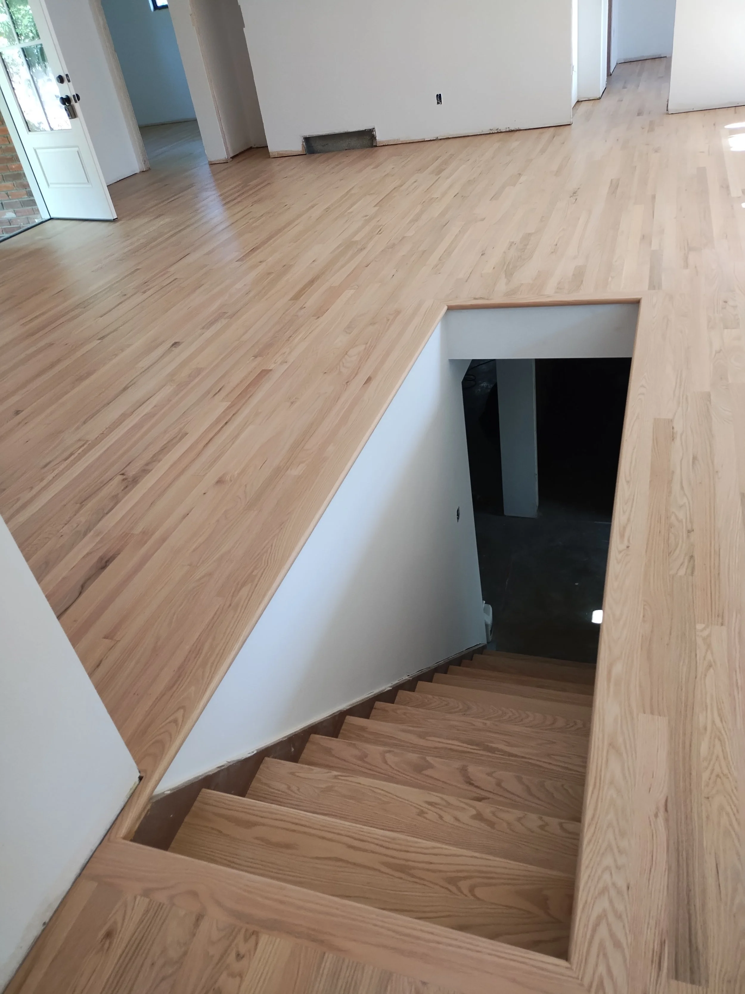 Interior of a house with light wood flooring, a staircase with a hole in the floor, and doorways leading to rooms. The flooring is partially finished.