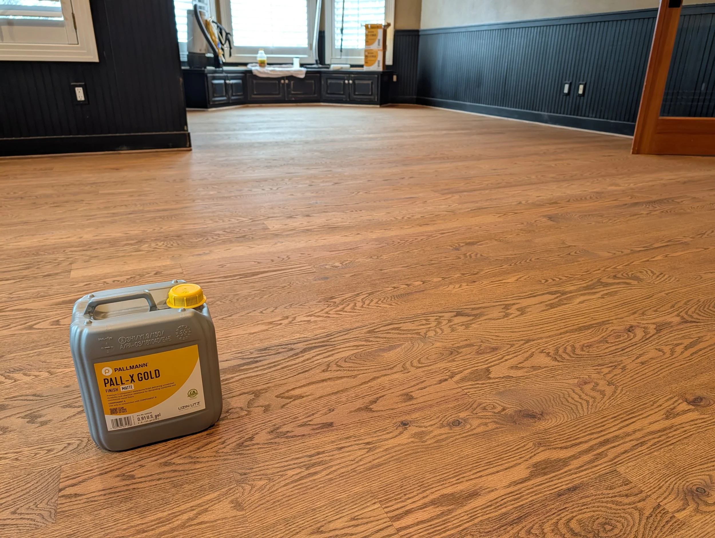 An empty room with dark wood paneling on the walls and a light wood floor. A can of Pallmann Pall-X Gold wood finish is in the foreground, and there is a small part of a yellow object near it. The room has a window with blinds and a black countertop 