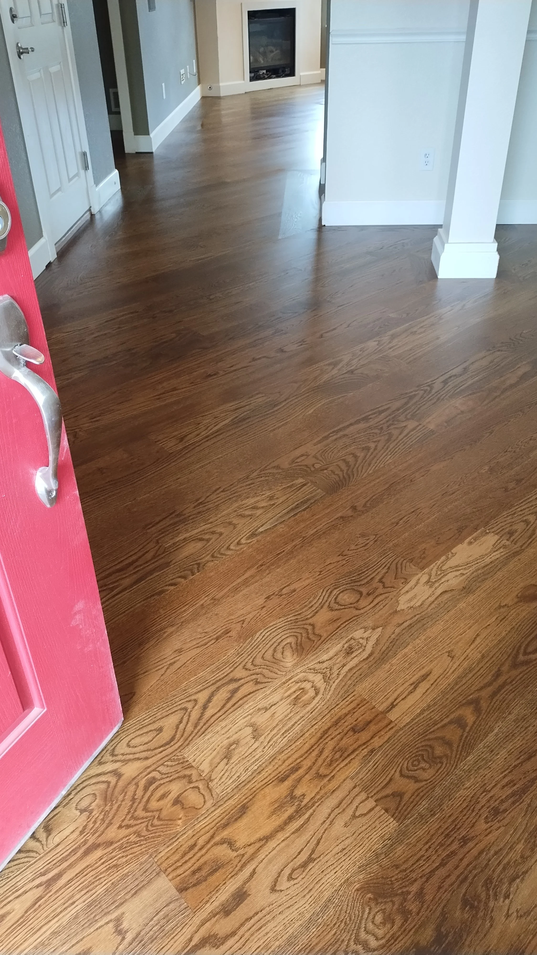 View of a hardwood floor and part of a pink door with a silver handle, and a hallway leading to a living room with a fireplace, with light-colored walls and baseboards.