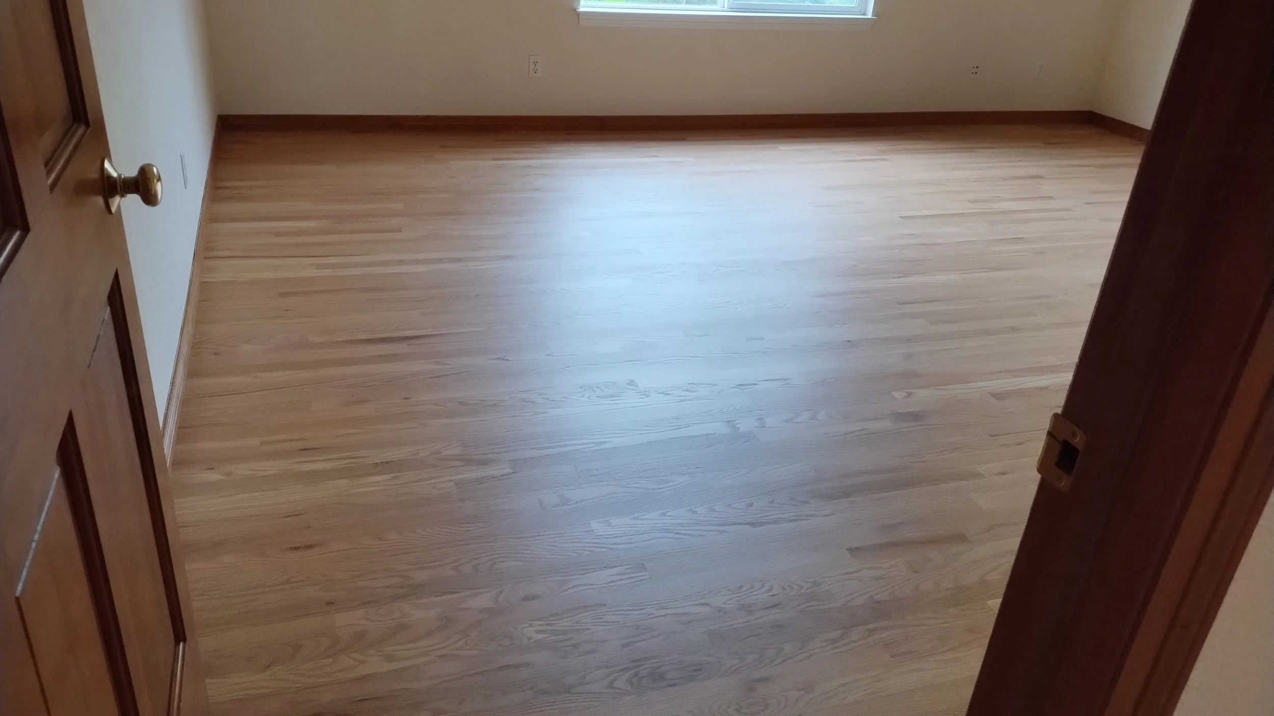 Empty room with light wood flooring, white walls, a single window, and a partially open wooden door.