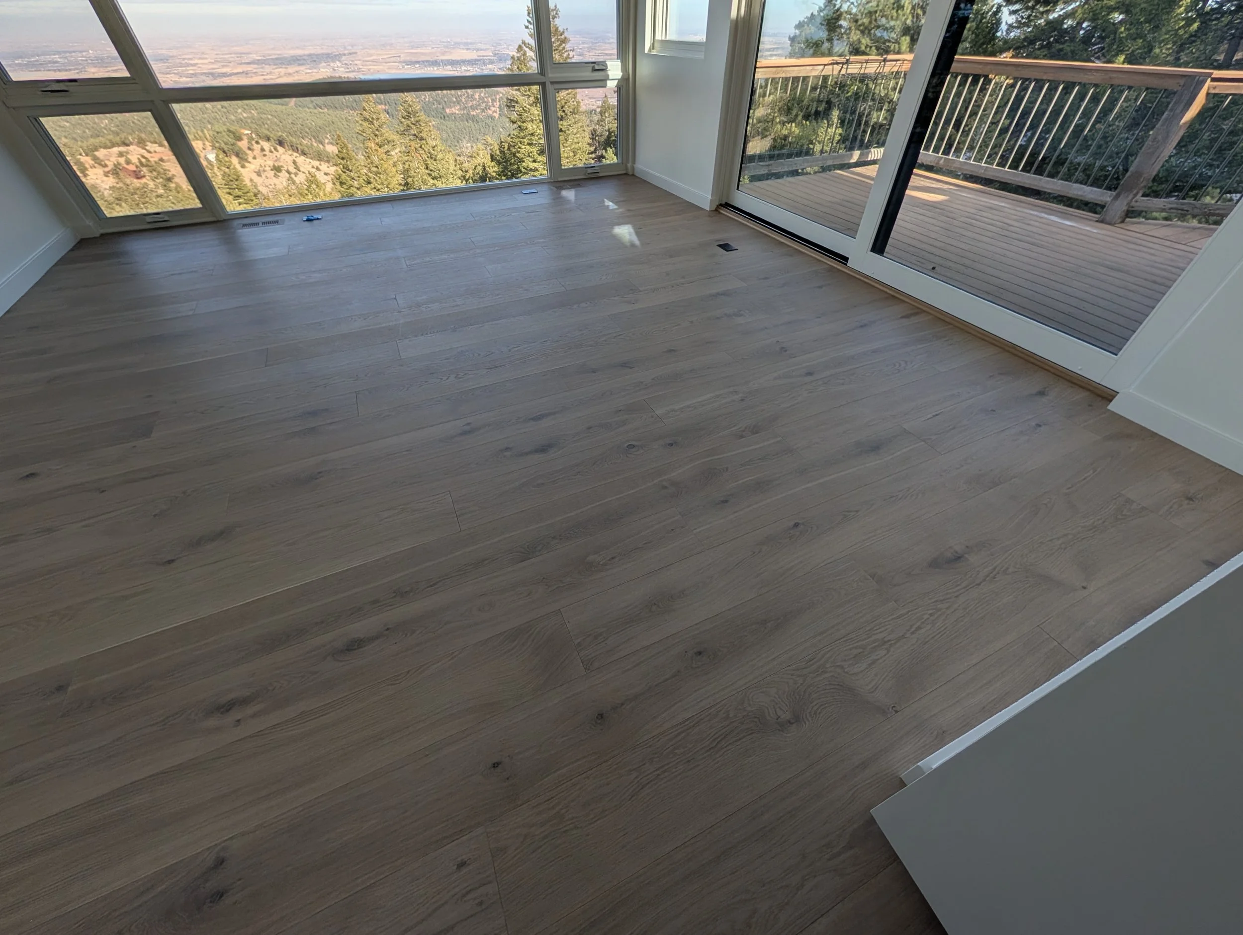 A room with light-colored wooden flooring, large windows showcasing a view of a forested mountainside and distant landscape, and a sliding glass door leading to a wooden balcony railing.
