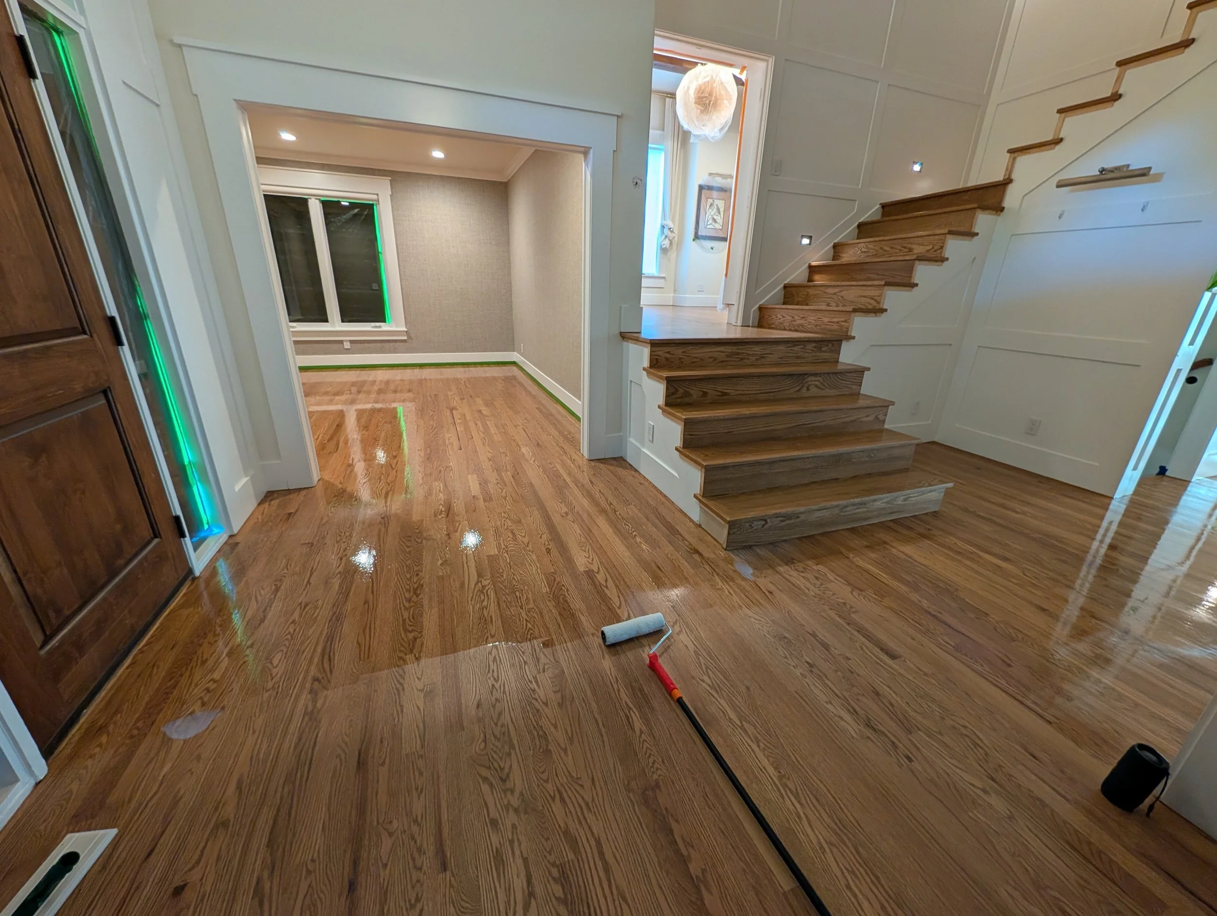 Interior of a house under renovation with hardwood floors, a staircase with wooden steps, a window, and a hanging paper lantern. A paint roller and paint tray are on the floor.