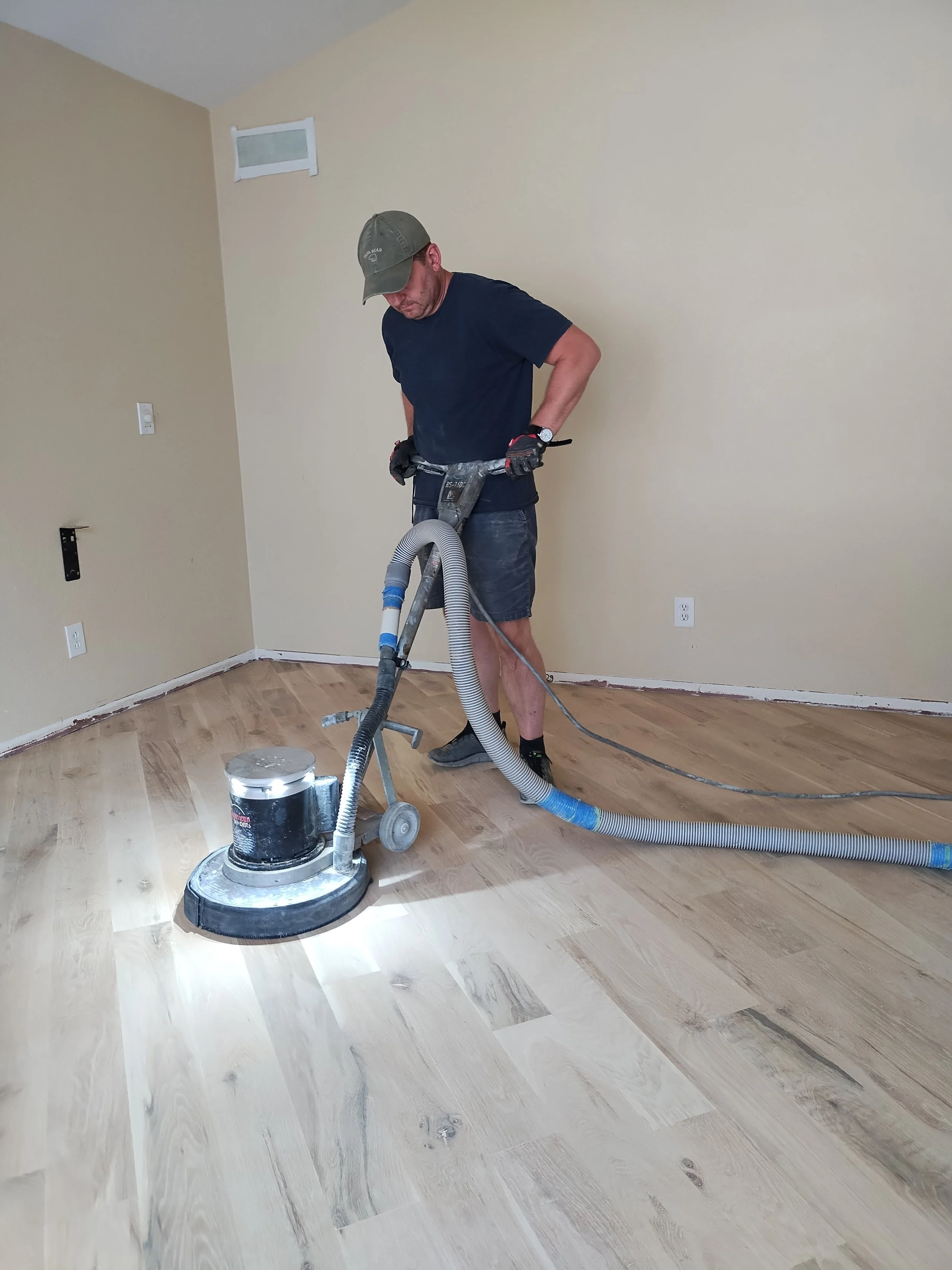 A man operating a floor sanding machine in a room with light-colored wooden flooring, beige walls, and a ceiling with a small window.
