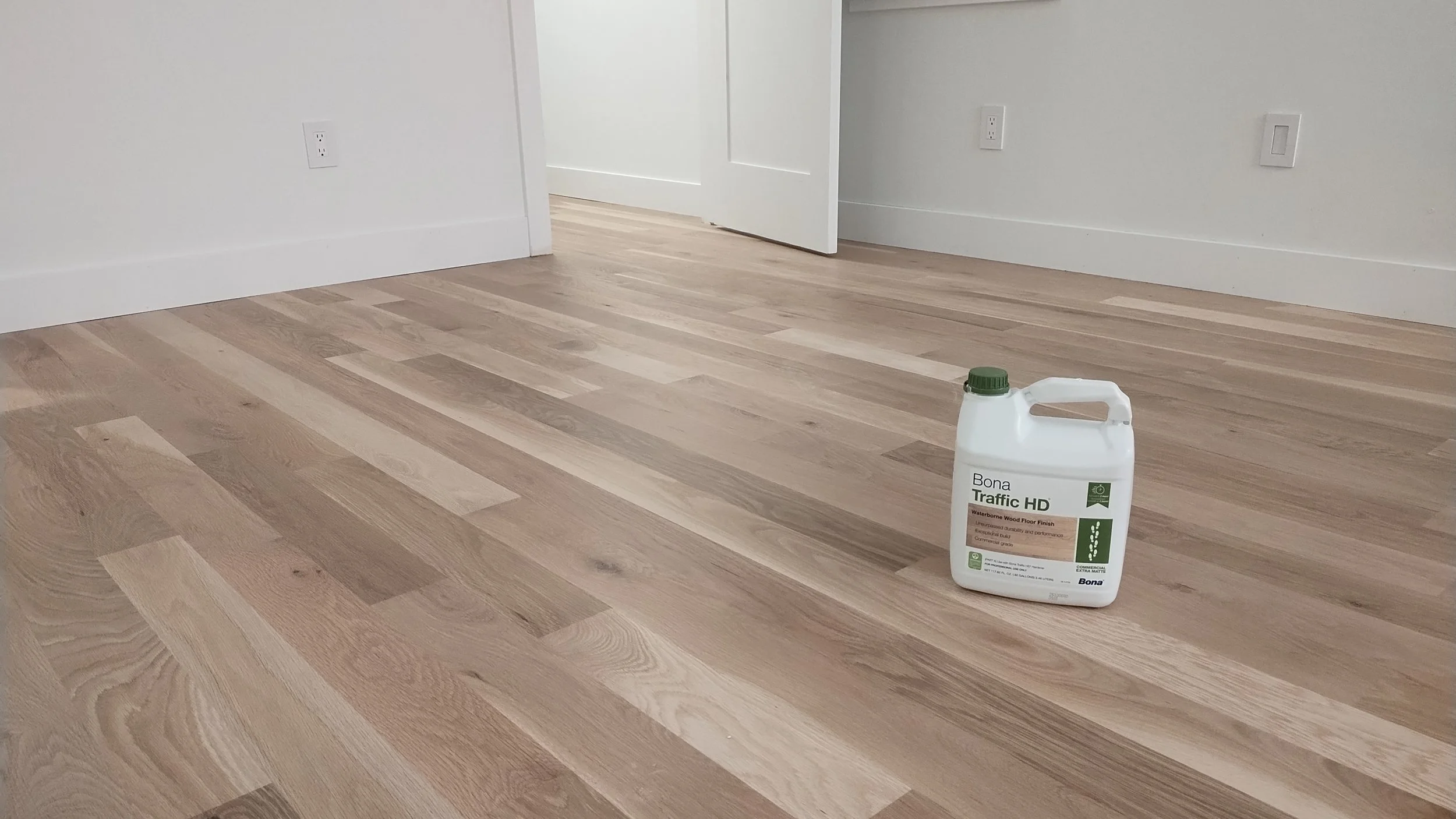 Room with light wood flooring and white walls, with an open door and a container of Bona Traffic HD wood floor finish on the floor.