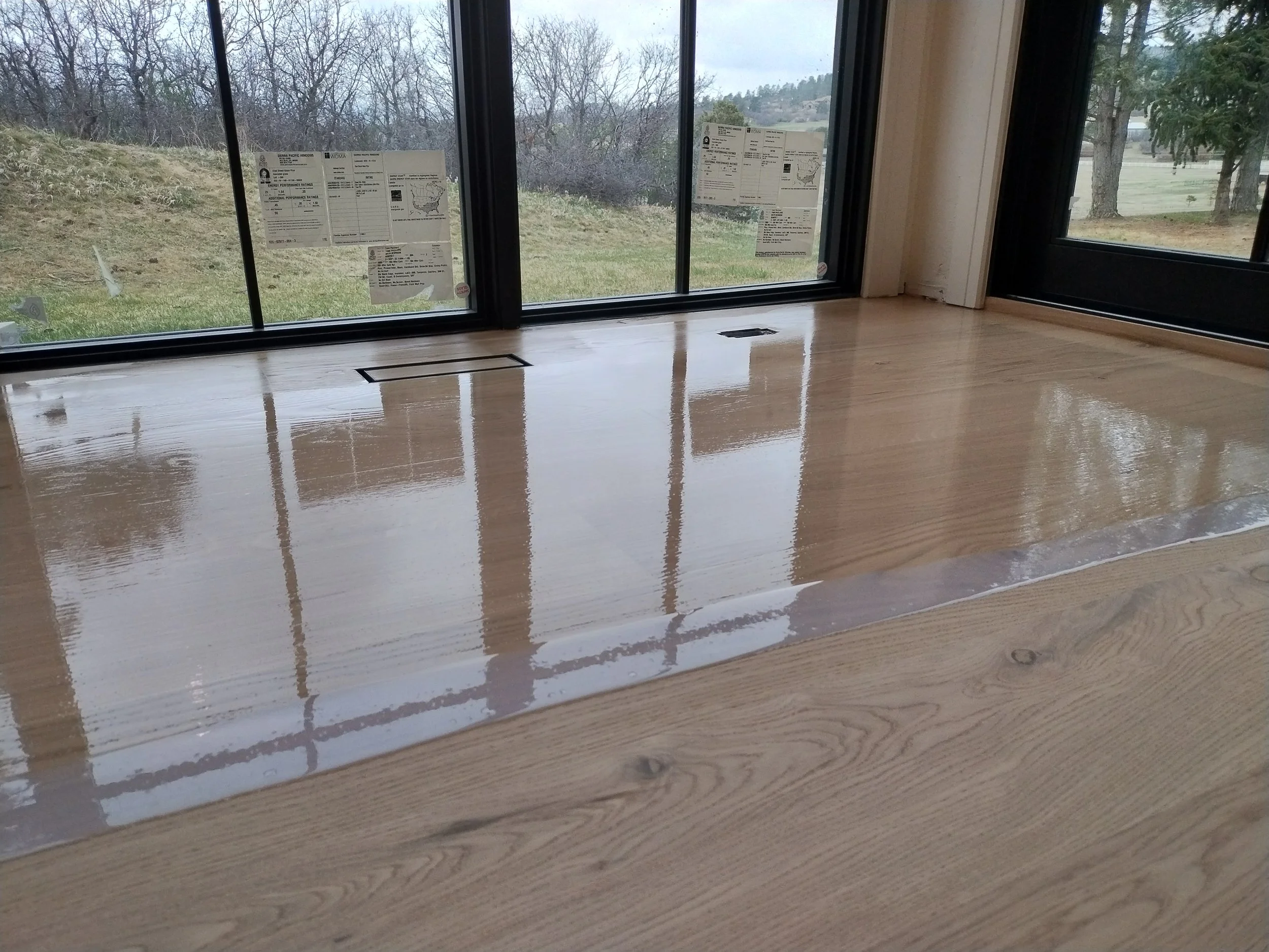 Interior view of a room with a window showing a cloudy outdoor landscape with trees and grass. The window has stickers with information, possibly related to the glass or building specifications. The floor appears to be freshly polished wood, reflecti