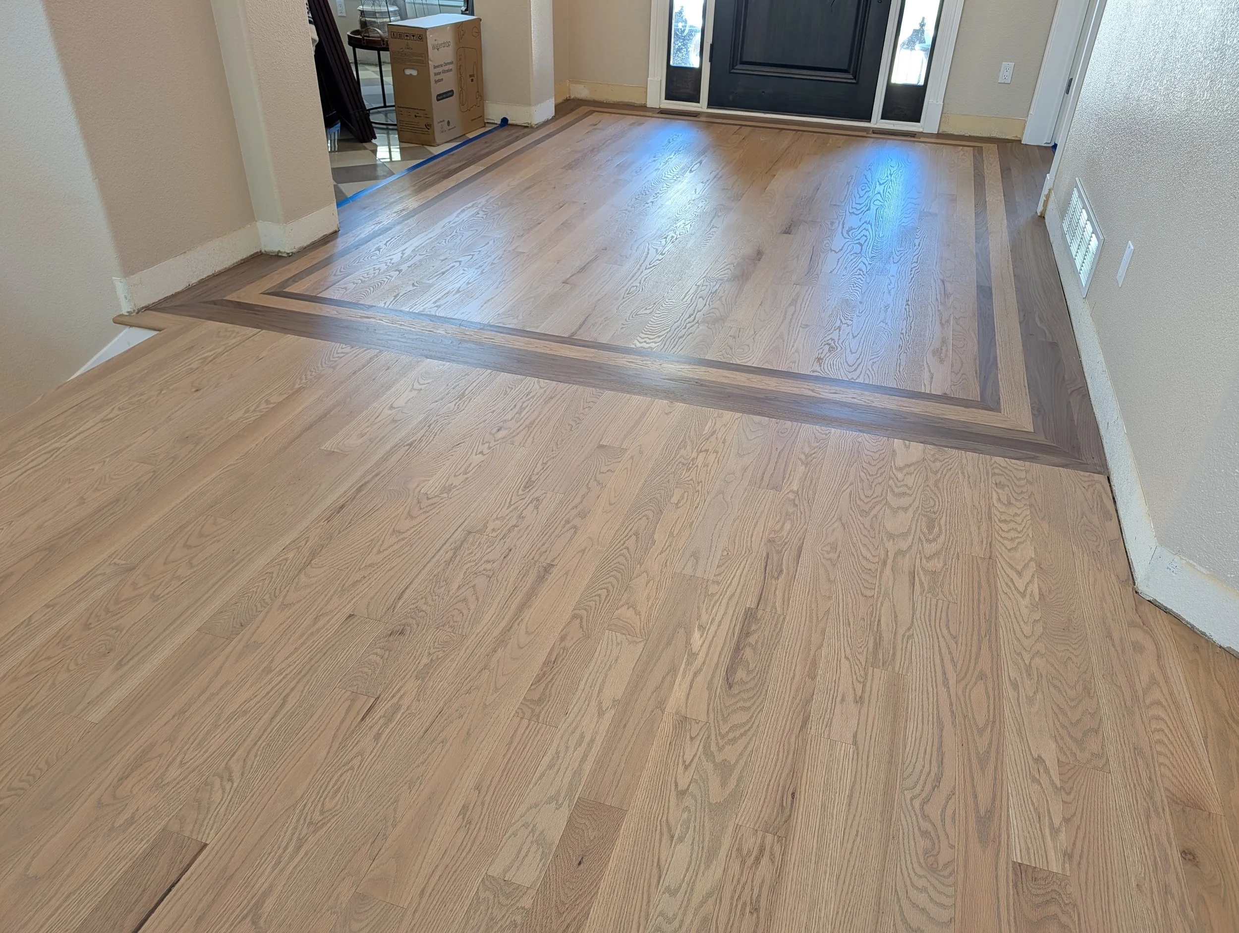 A wooden floor with a decorative border next to a front door and hallway. A cardboard box is visible near the entrance.