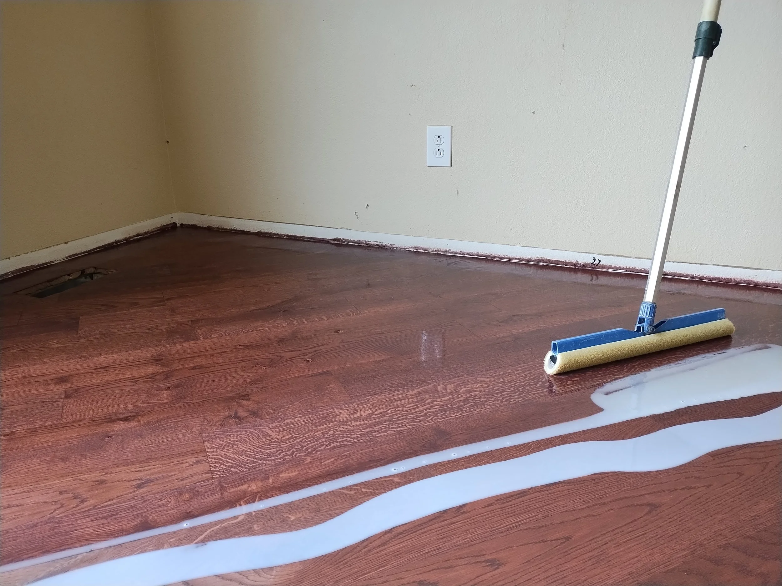 Empty room with wooden floor, beige wall with electrical outlet, patch of water on the floor, and a paint roller in the corner with paint spilled around.