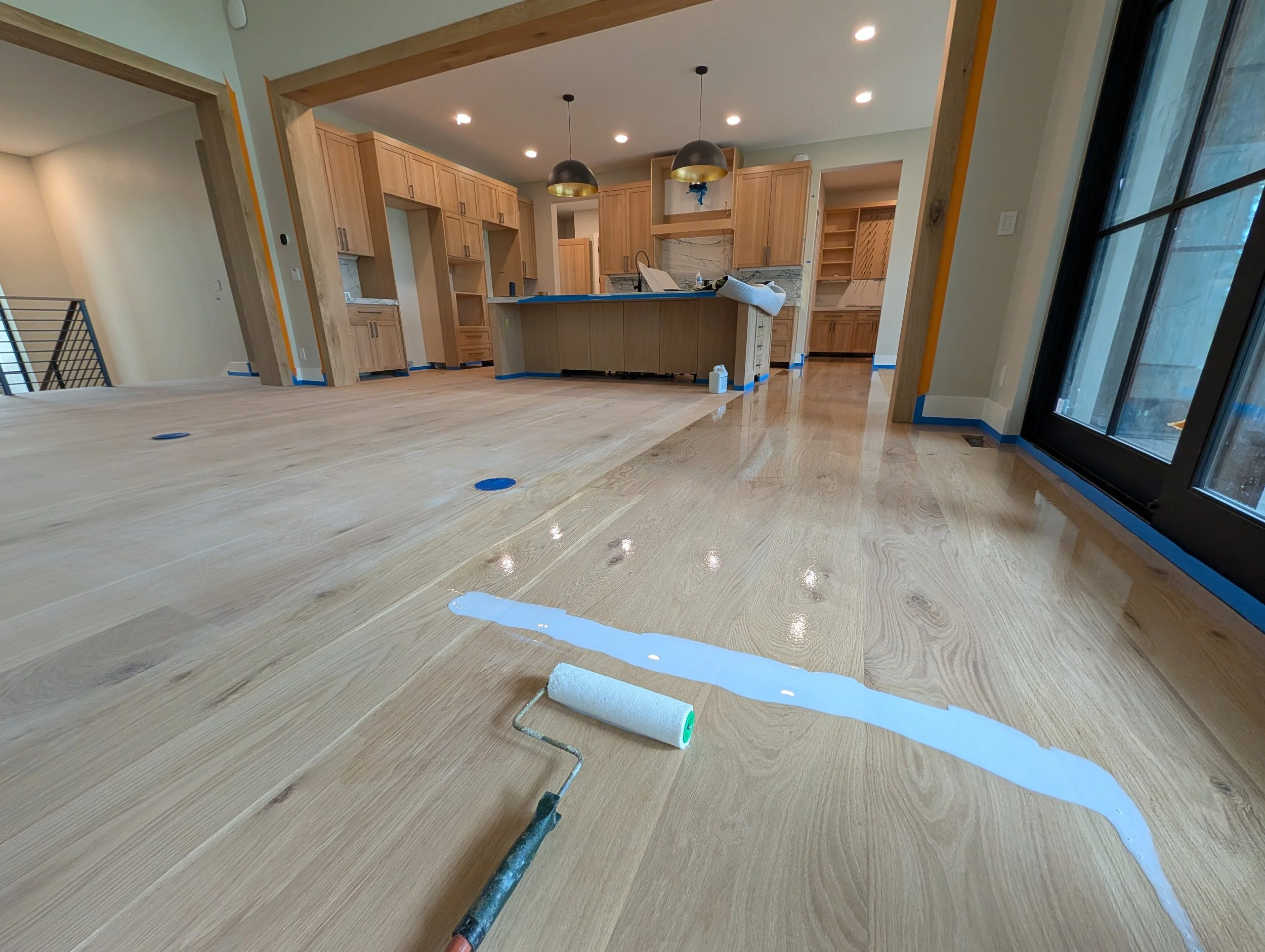 Interior of a house under construction or renovation featuring a kitchen with wooden cabinetry and countertops, large windows, and a floor covered with a glossy finish. Painting supplies and tools are visible on the floor.