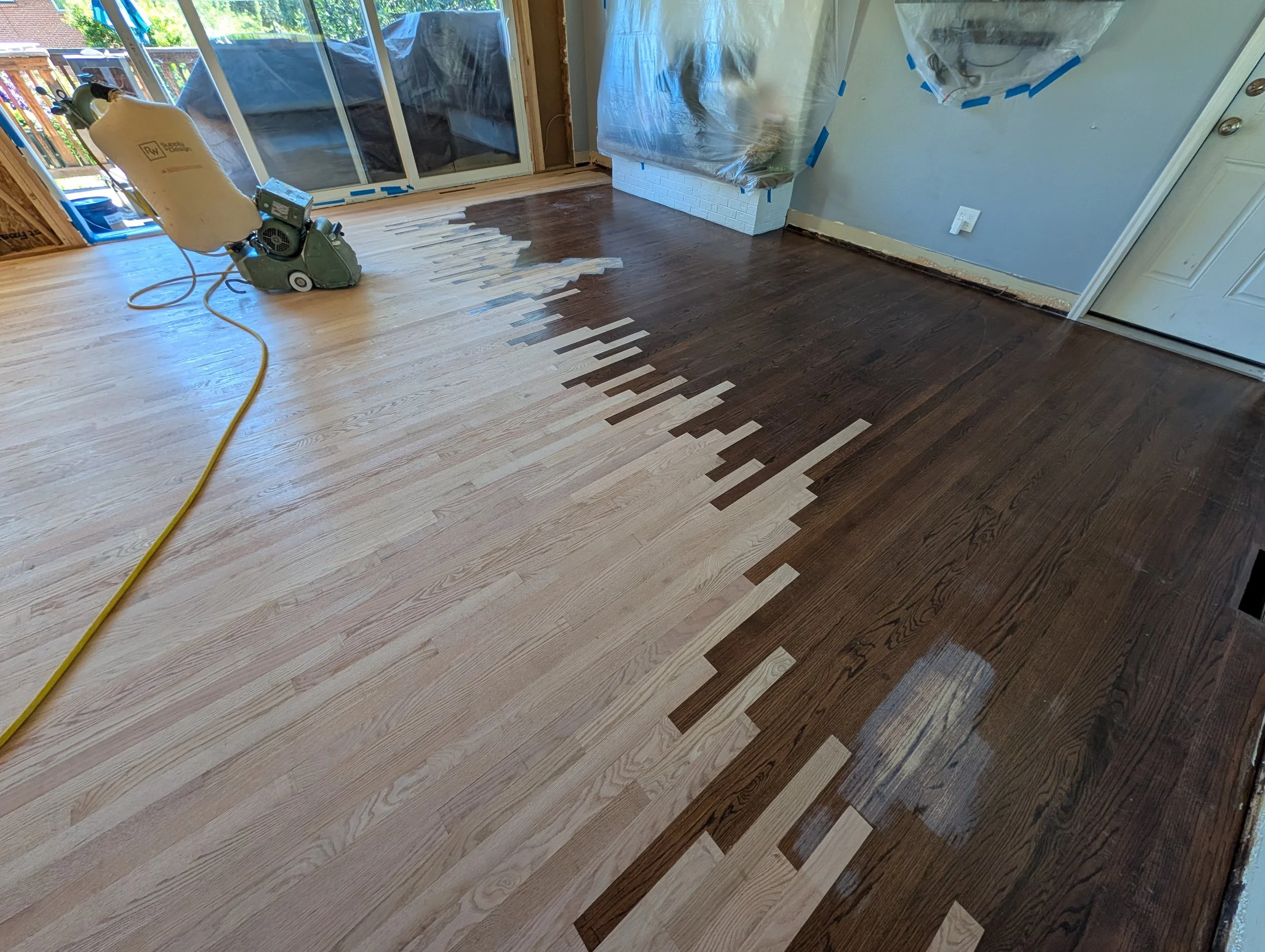 A room with hardwood flooring in the process of being refinished, with a floor sander and an extension cord visible. The room has large glass sliding doors leading to an outdoor deck, and the walls are partially covered with plastic sheeting for protection during renovation.
