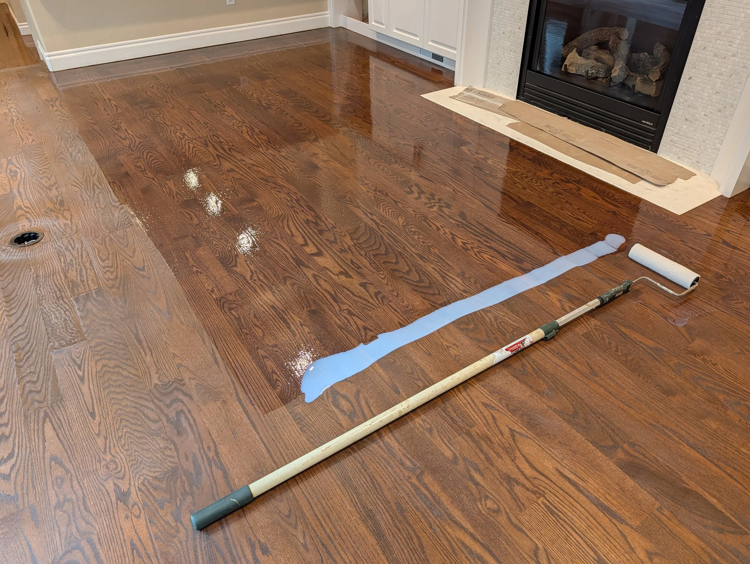 Refinished hardwood floor next to a fireplace with area protected by cardboard, and a paint roller nearby, indicating ongoing renovation work.