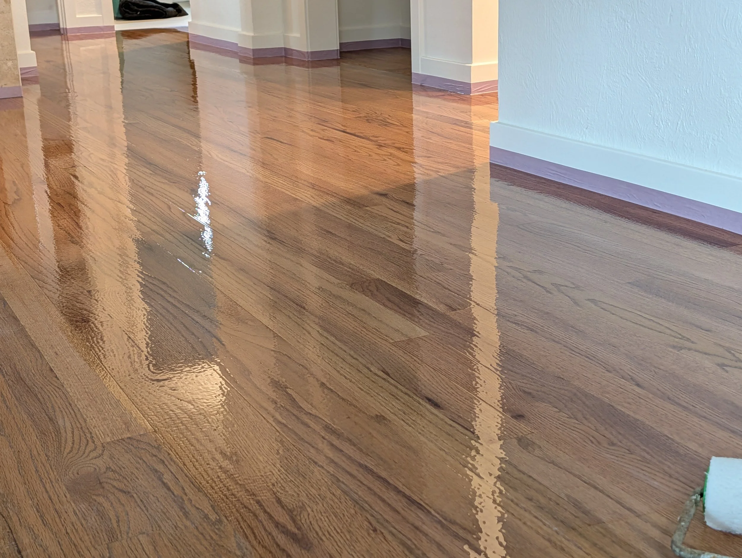 Close-up photograph of a polished hardwood floor with a reflective surface, showing a section of a room's corner with white baseboards, white walls, and a doorway in the background.