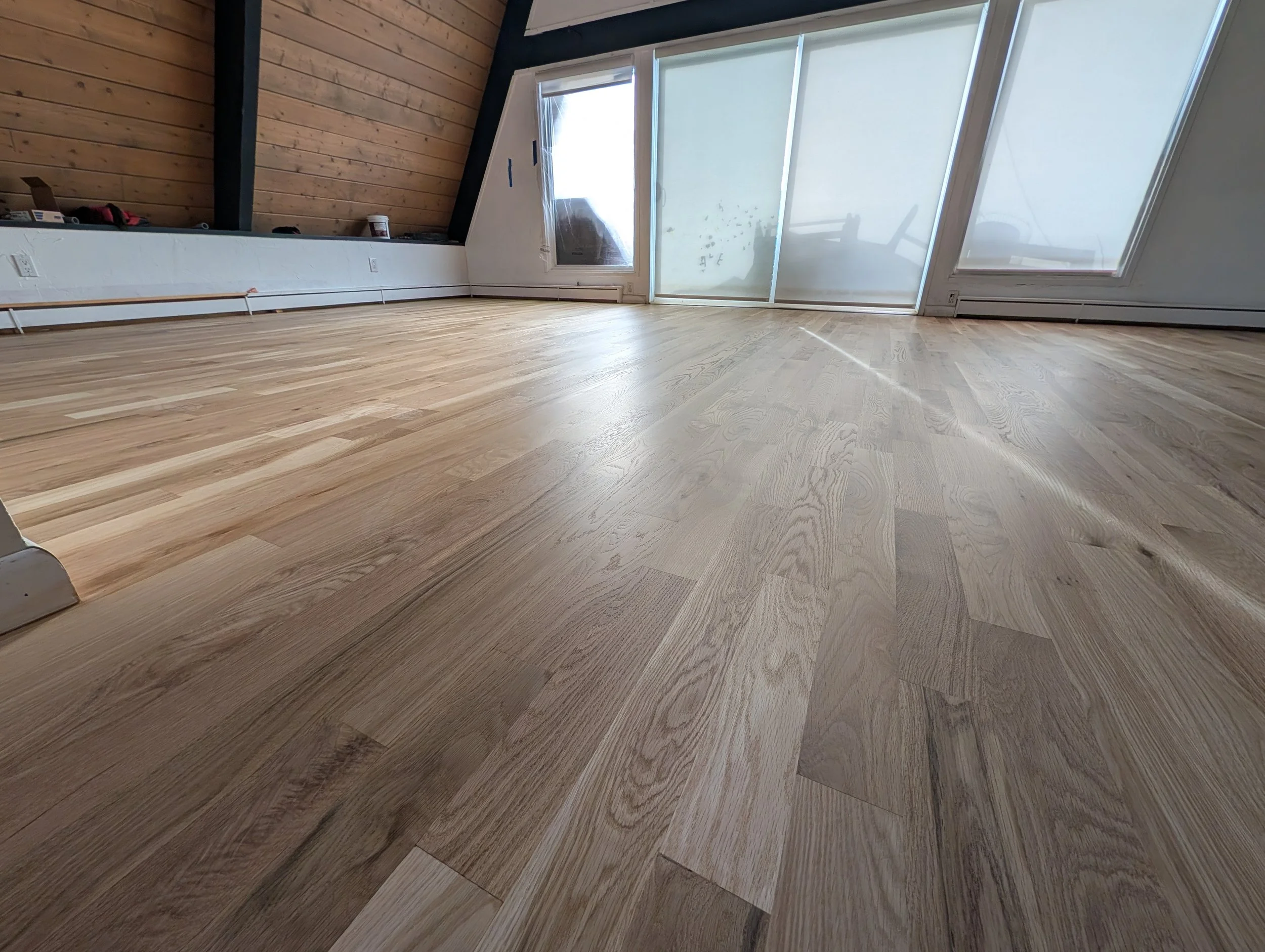 Empty room with hardwood floors, wooden wall paneling, large windows with blinds, and some scattered items.