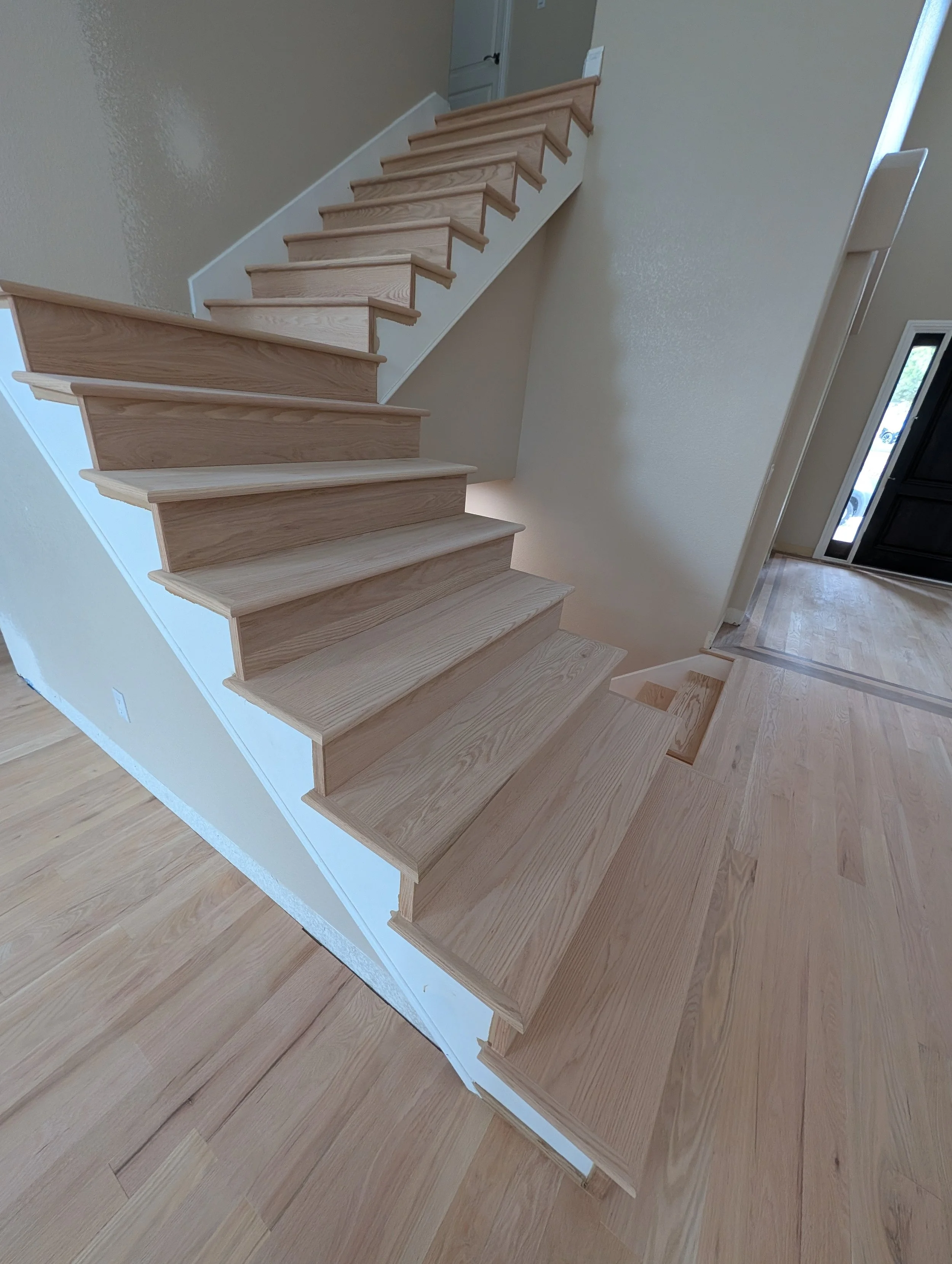 Interior view of wooden staircase with light-colored wood steps and white sides, leading to a door at the top, with hardwood flooring and a beige wall.