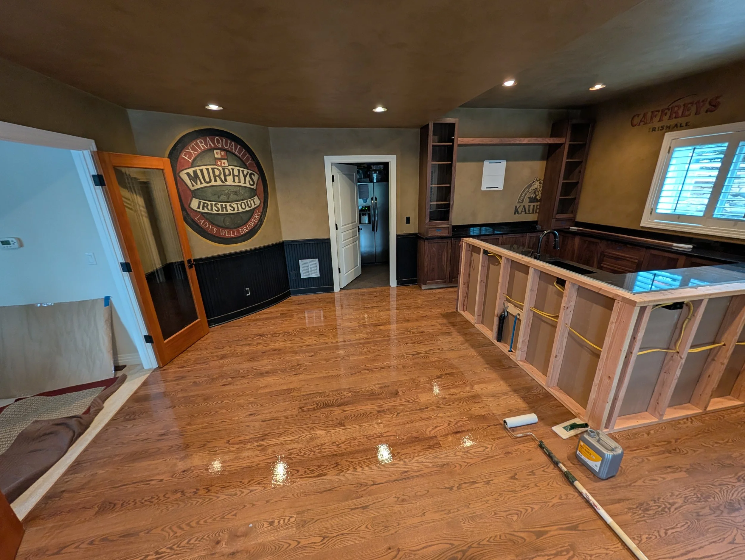 Interior of a room under renovation with unfinished kitchen cabinets, wooden flooring, and walls decorated with beer brand posters. Construction tools and supplies are scattered on the floor.