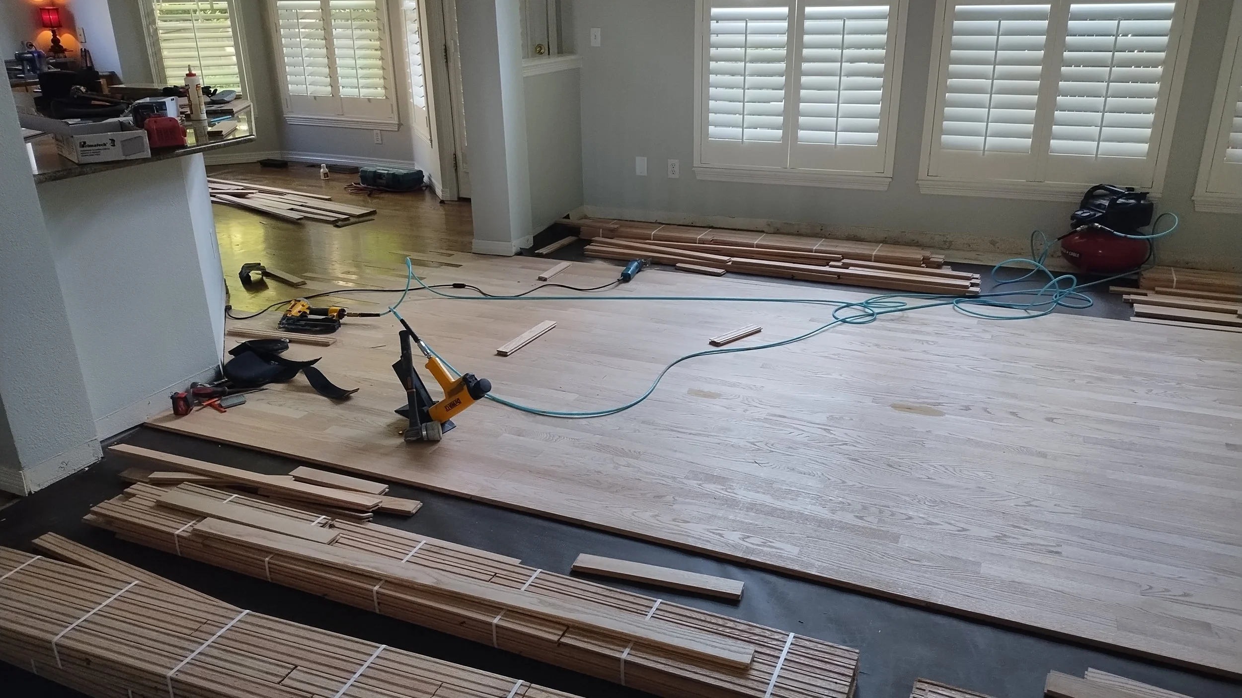 Living room with hardwood flooring under renovation, with stacks of wooden flooring planks, tools, and equipment scattered across the floor and near the wall.