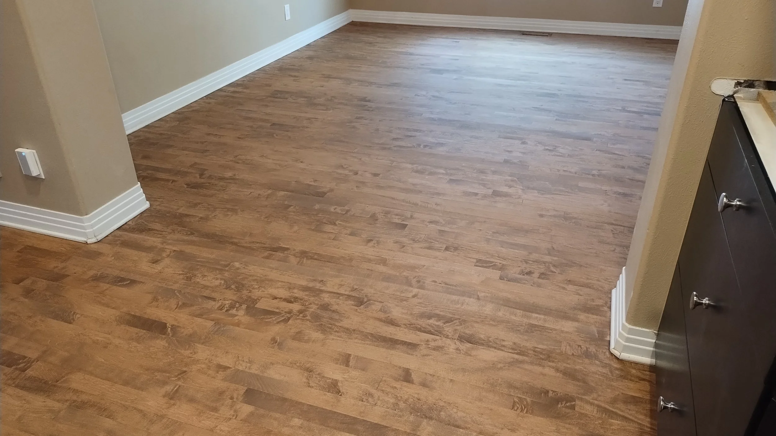 Empty room with hardwood floors, beige walls, white baseboards, and a small section of dark cabinetry on the right.