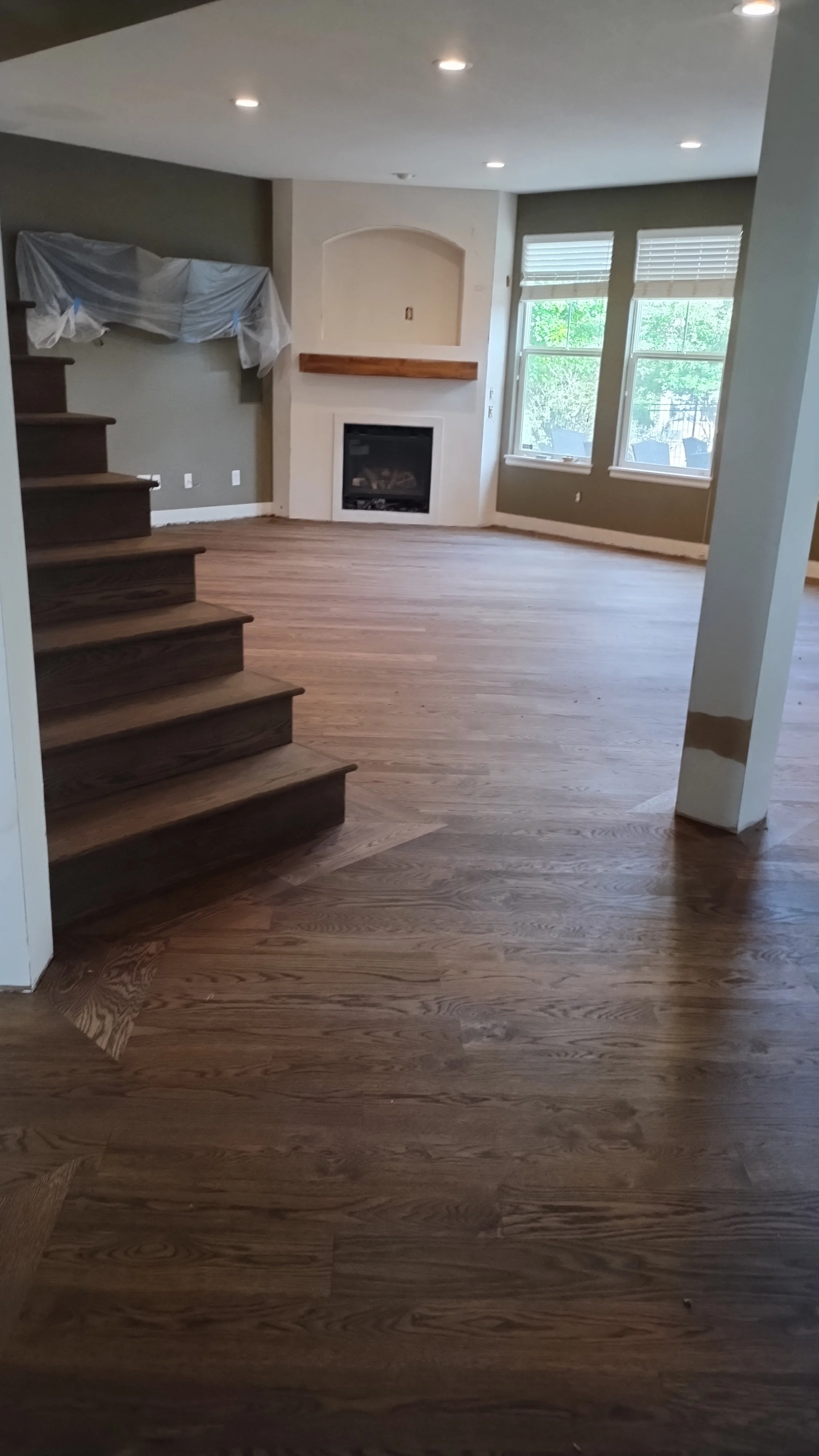 Inside a house, showing a living room with wooden flooring, a staircase on the left, a fireplace, and large windows letting in natural light.