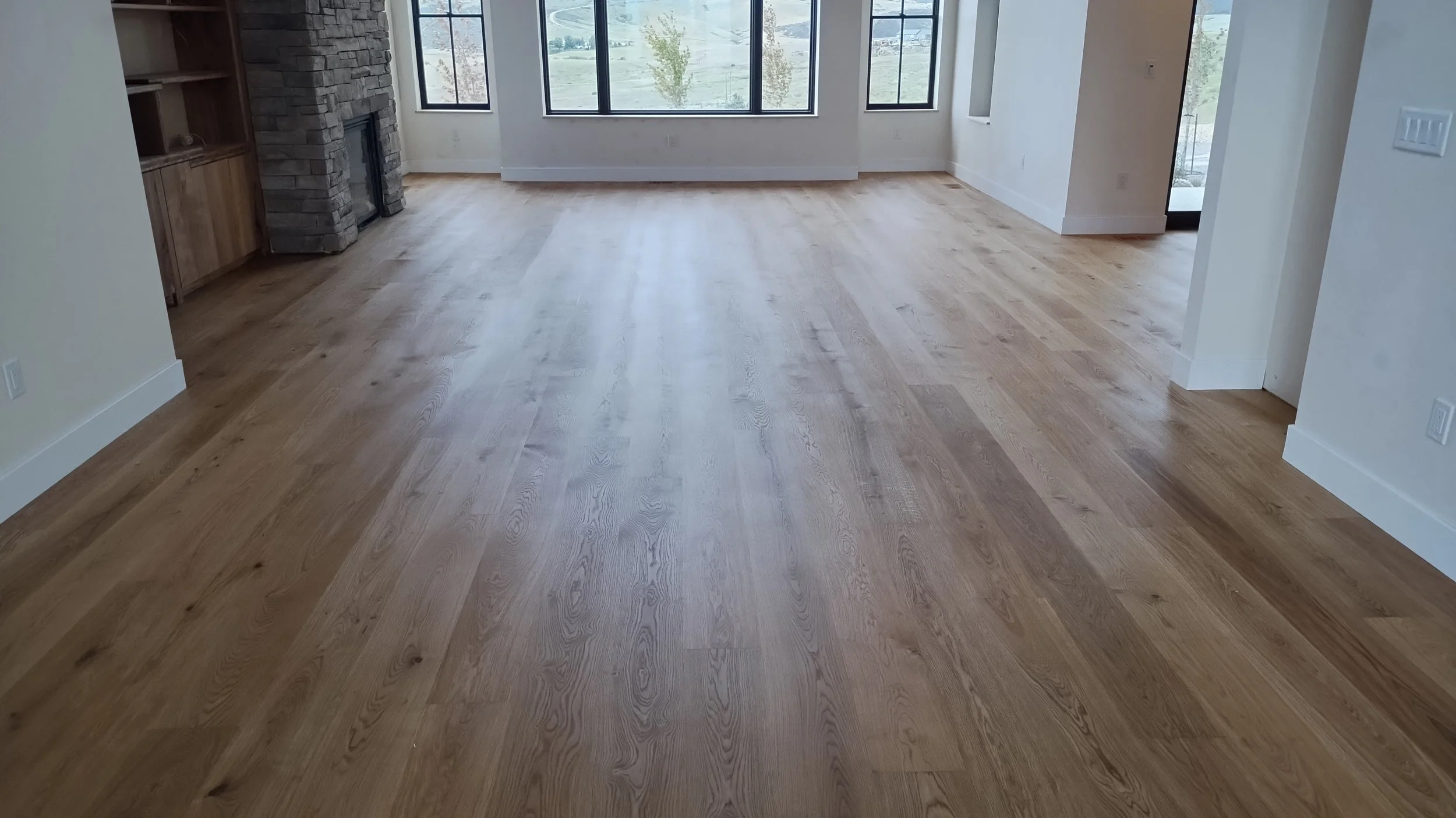 Empty living room with light hardwood floors, white walls, large front windows, and a stone fireplace.