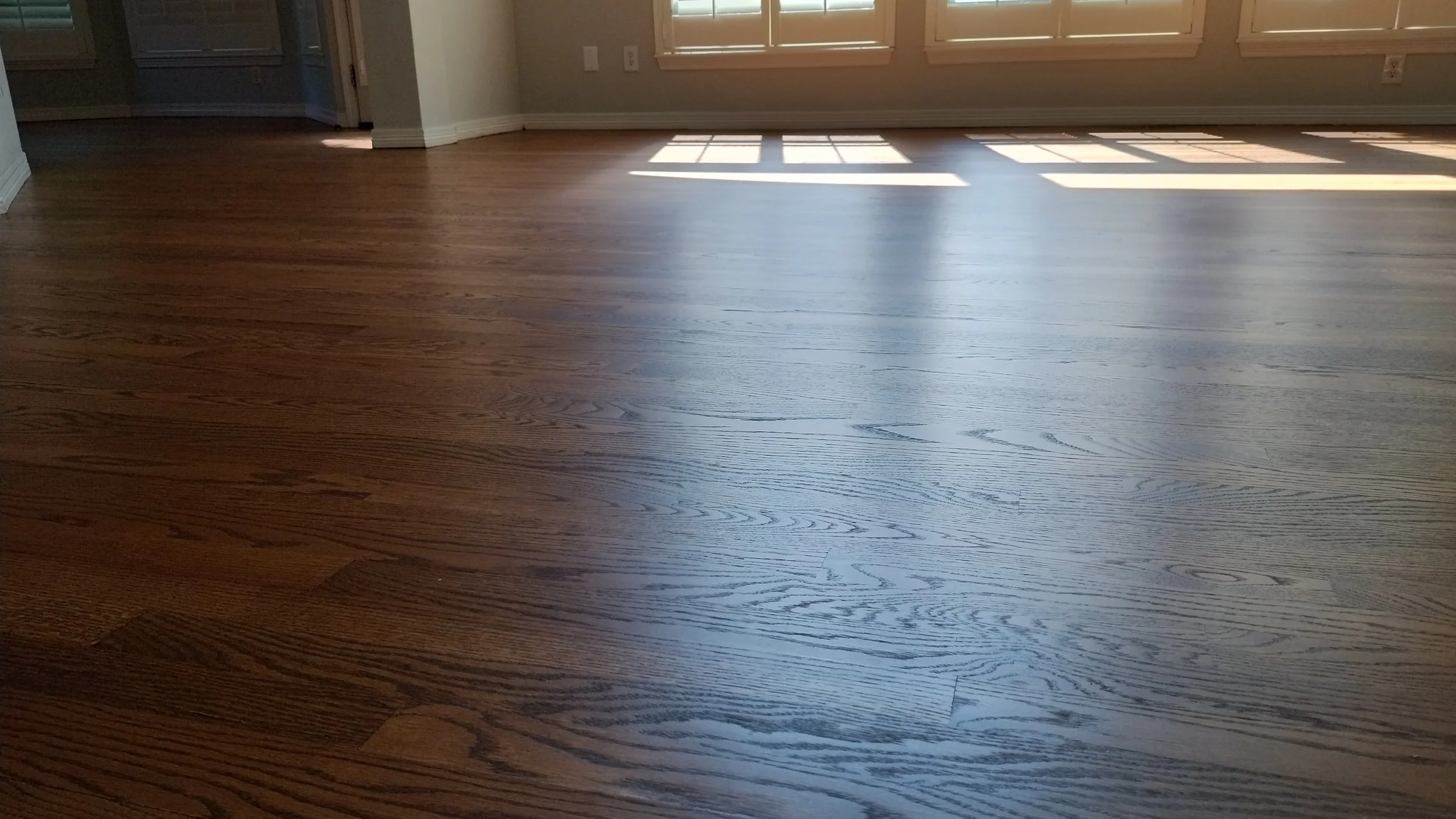 Freshly polished hardwood floor in a sunlit room with white window blinds, On the left side, a partial front hall with some accessible electrical outlets.