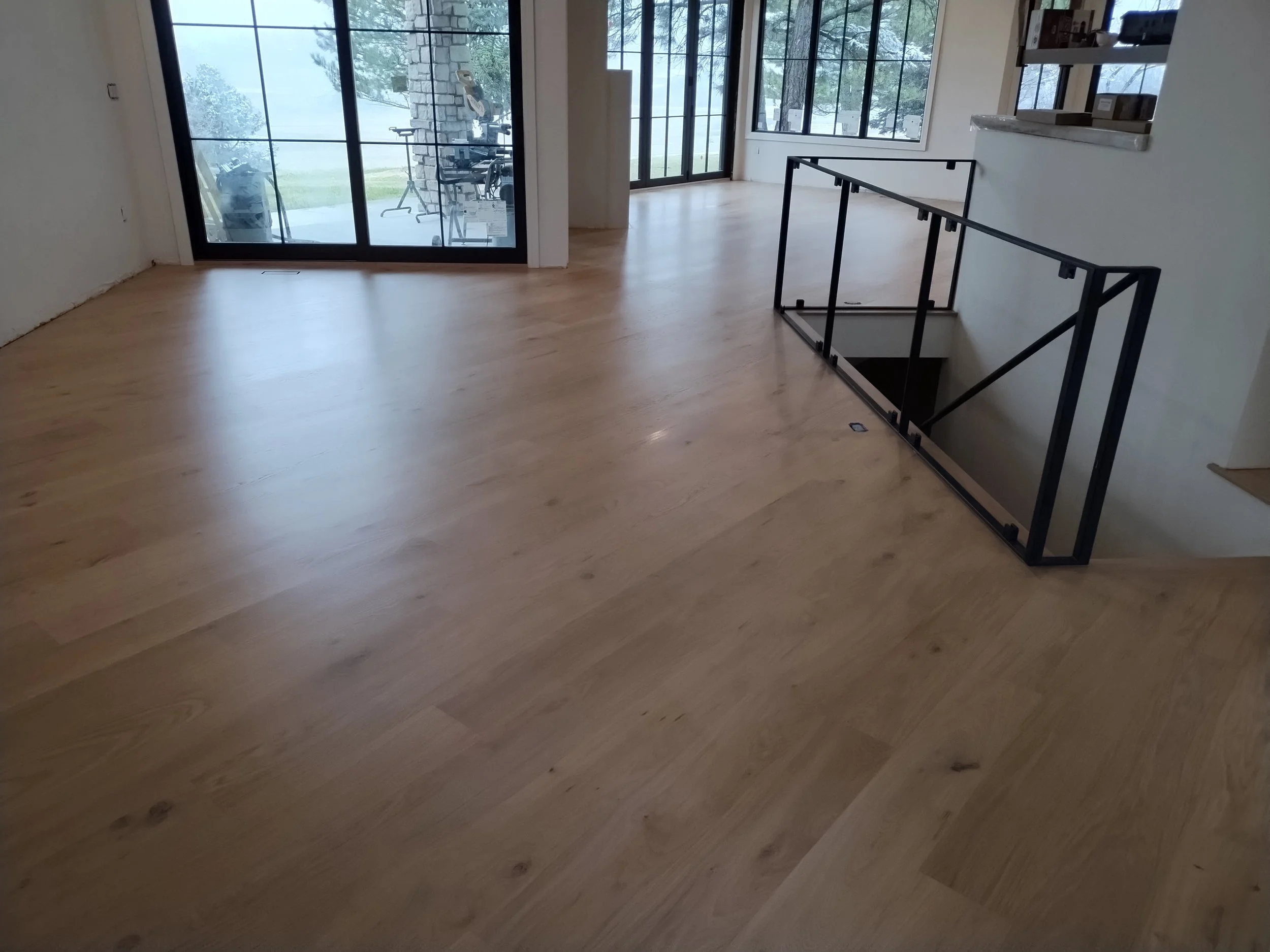 Empty room with light wood flooring, large windows, and a railing near the stairs. Outside, a patio with exercise equipment is visible through the windows.