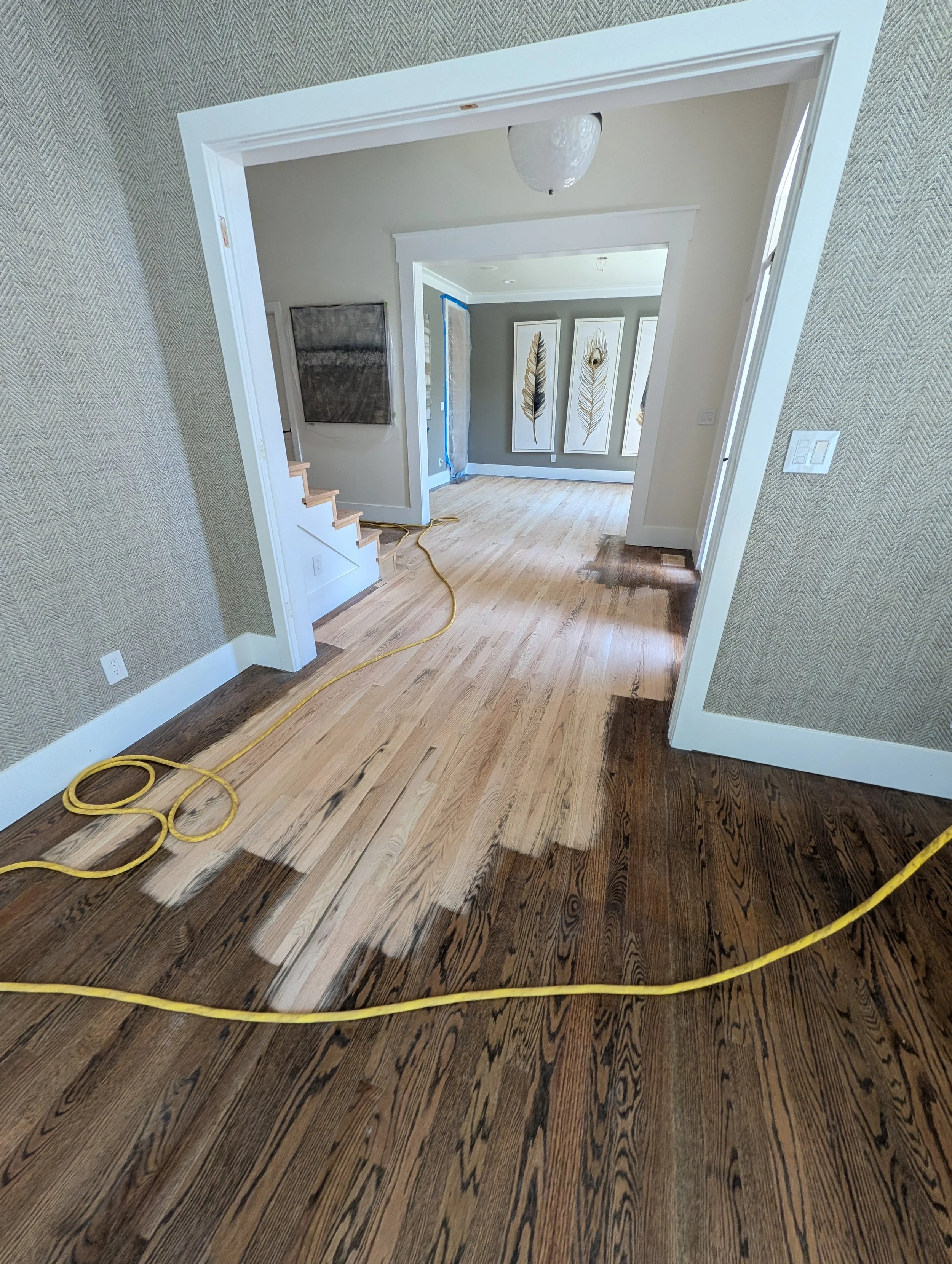 Interior room with hardwood flooring, beige and gray walls, decorative artwork, a staircase, and a hanging light fixture. Yellow extension cords are on the floor.