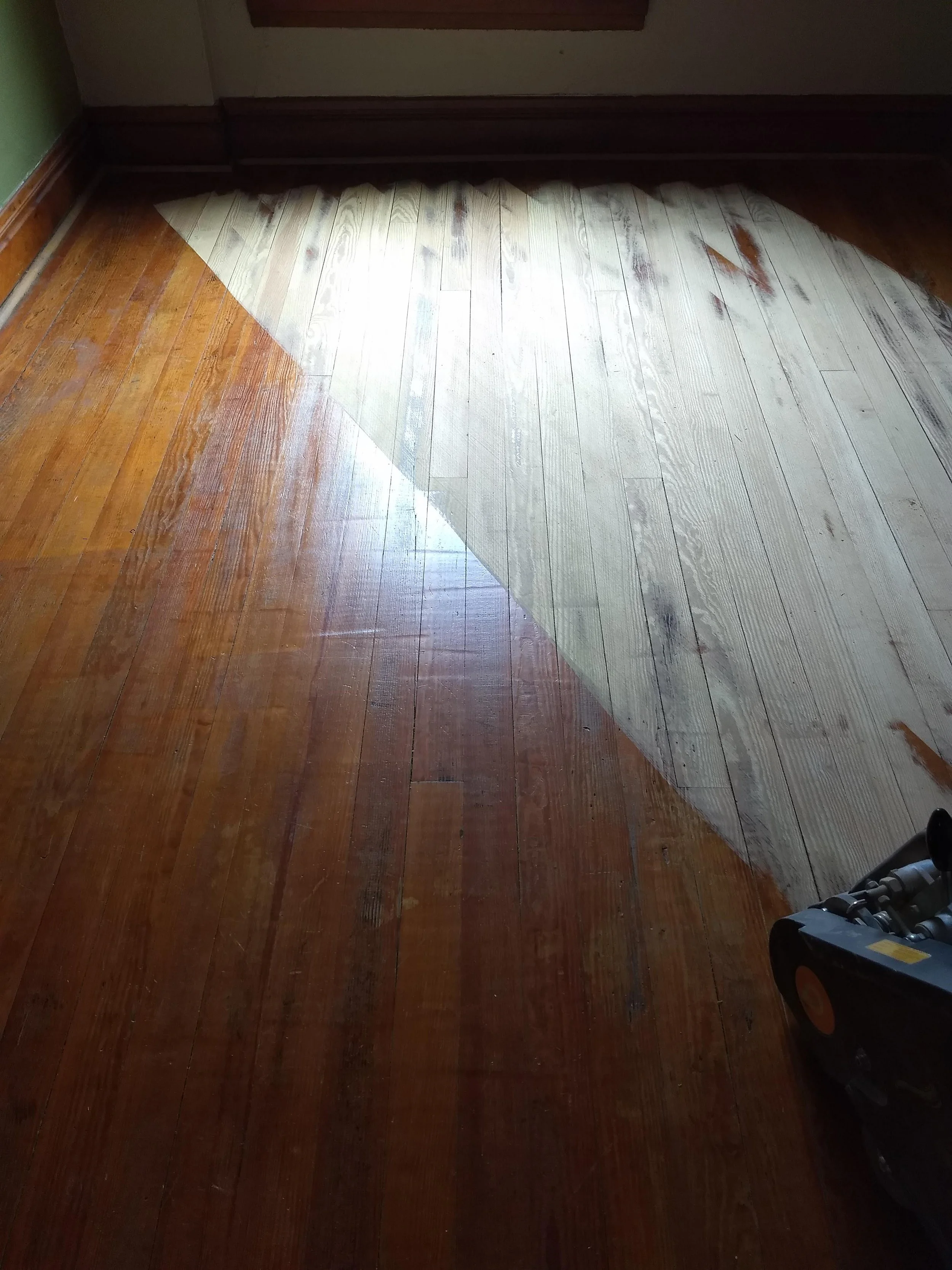 A hardwood floor in the process of being refinished, with part of it still in its original darker stain and part sanded and lighter in color, with woodworking tools visible in the corner.