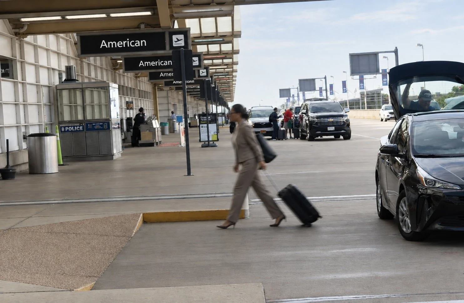 Passenger walking with a rolling suitcase hastly near the American Airlines section at an airport curbside. Other vehicles and people in the background, with a police bin, trash cans, and signage visible.