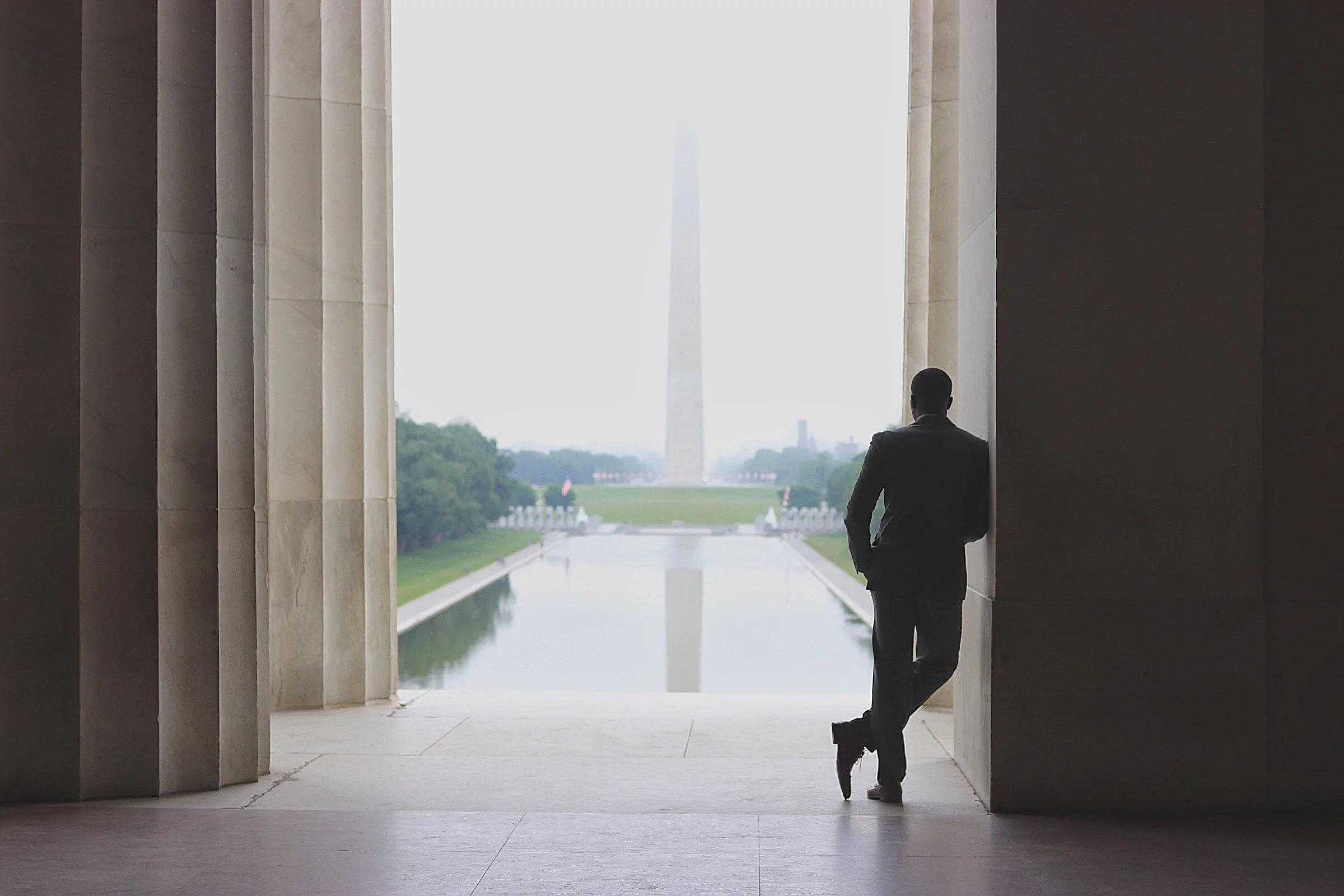 A man in a suit standing and leaning against a large open way column of the Lincoln Memorial, looking out at the Washington, D.C. monument across the Reflecting Pool.