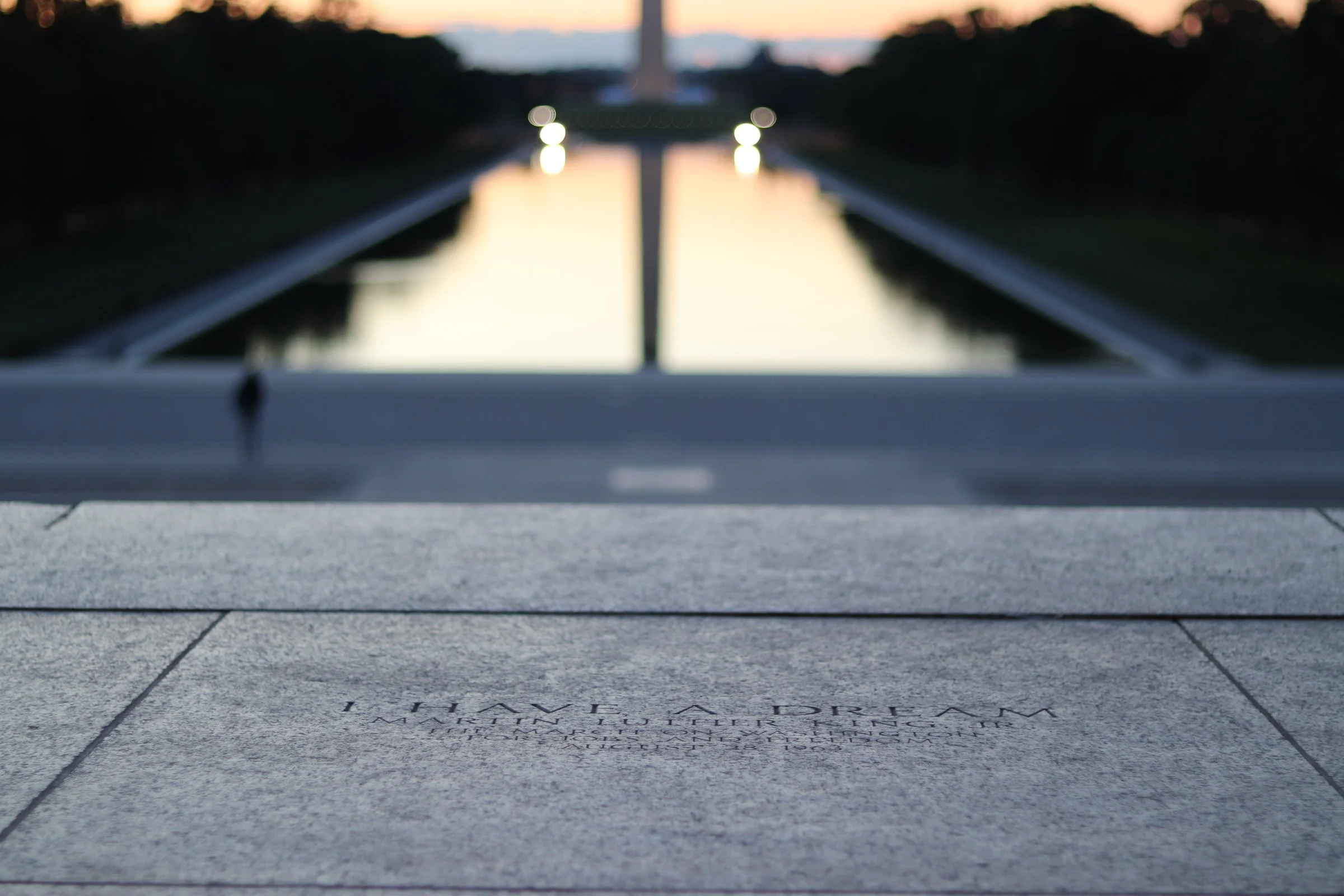View of the Lincoln Memorial Reflecting Pool at sunset with the Washington Monument in the distance, reflection of the sunset on the pool, and a blurred silhouette of trees on either side.