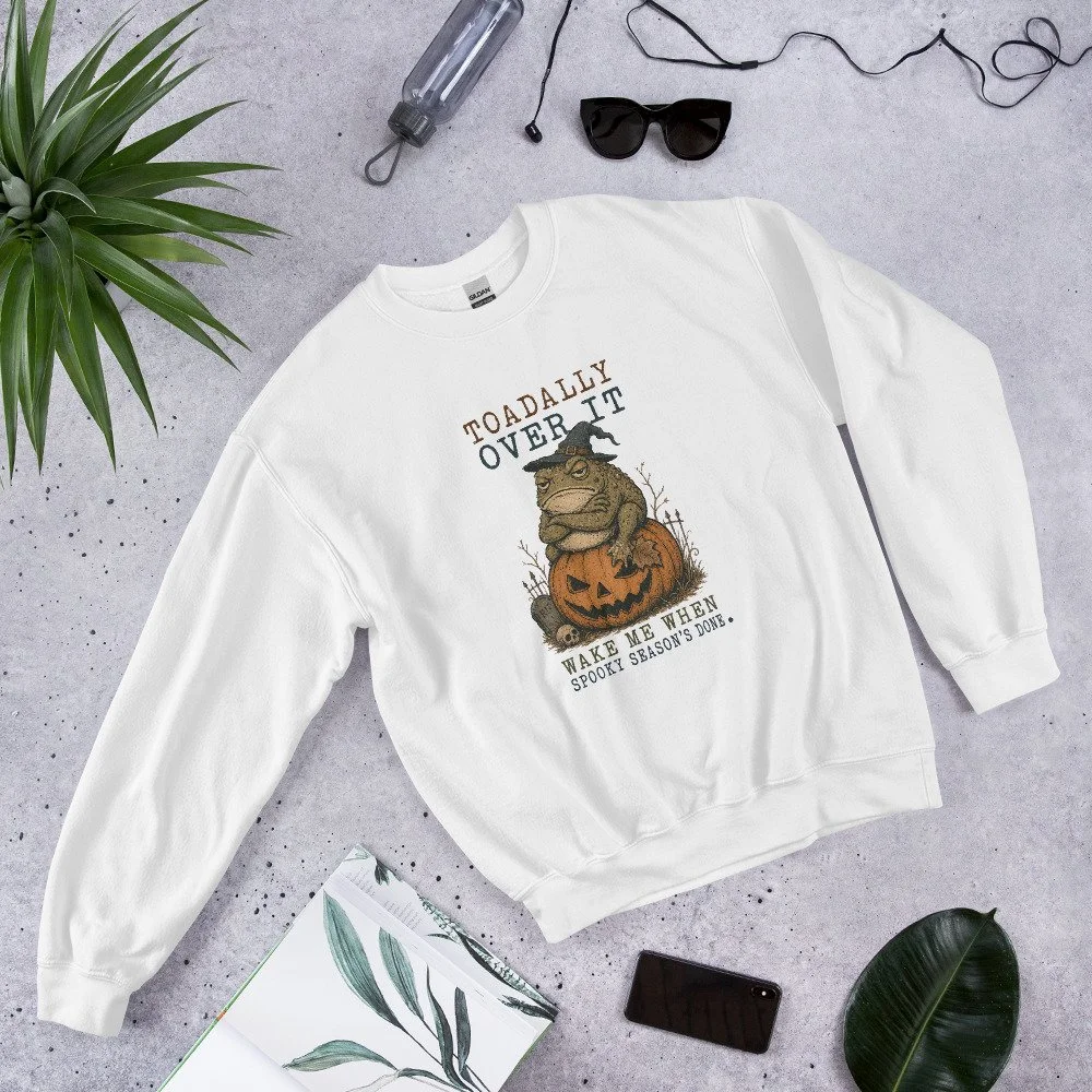 A white sweatshirt with a Halloween-themed graphic and text, surrounded by sunglasses, a water bottle, a plant, a notebook, a smartphone, and a large leaf, on a gray surface.