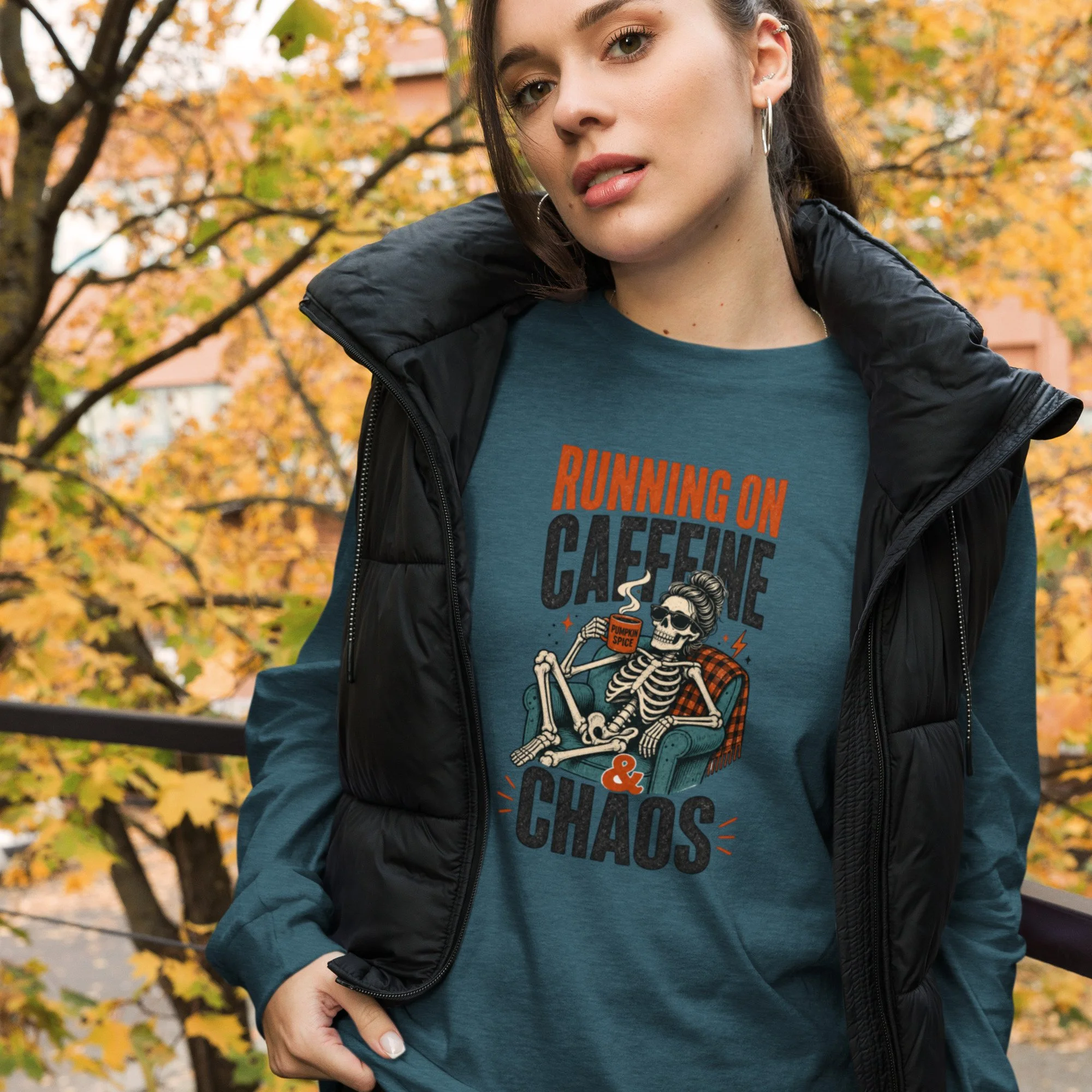 Close-up of a young woman with brown hair, wearing a black jacket and a teal T-shirt that has a skeleton graphic reading 'Running on Coffee & Chaos' printed on it, standing outdoors among fall trees with yellow and orange leaves.