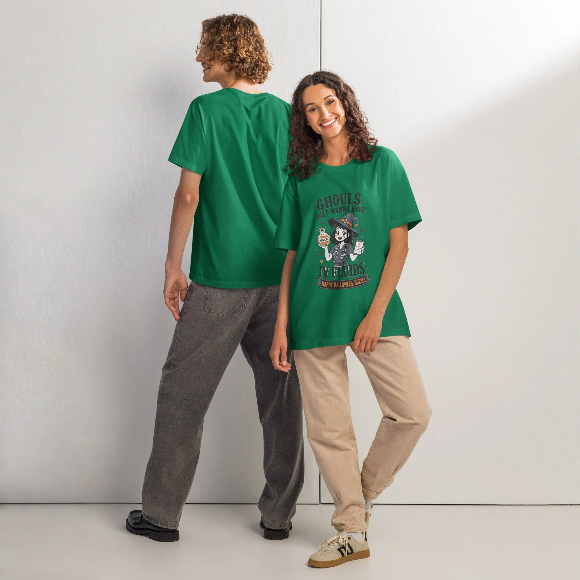 Two people standing back-to-back in front of a white wall, one with long curly hair wearing a green T-shirt, and the other with short curly hair wearing a matching green T-shirt with Halloween-themed graphics, and beige pants.