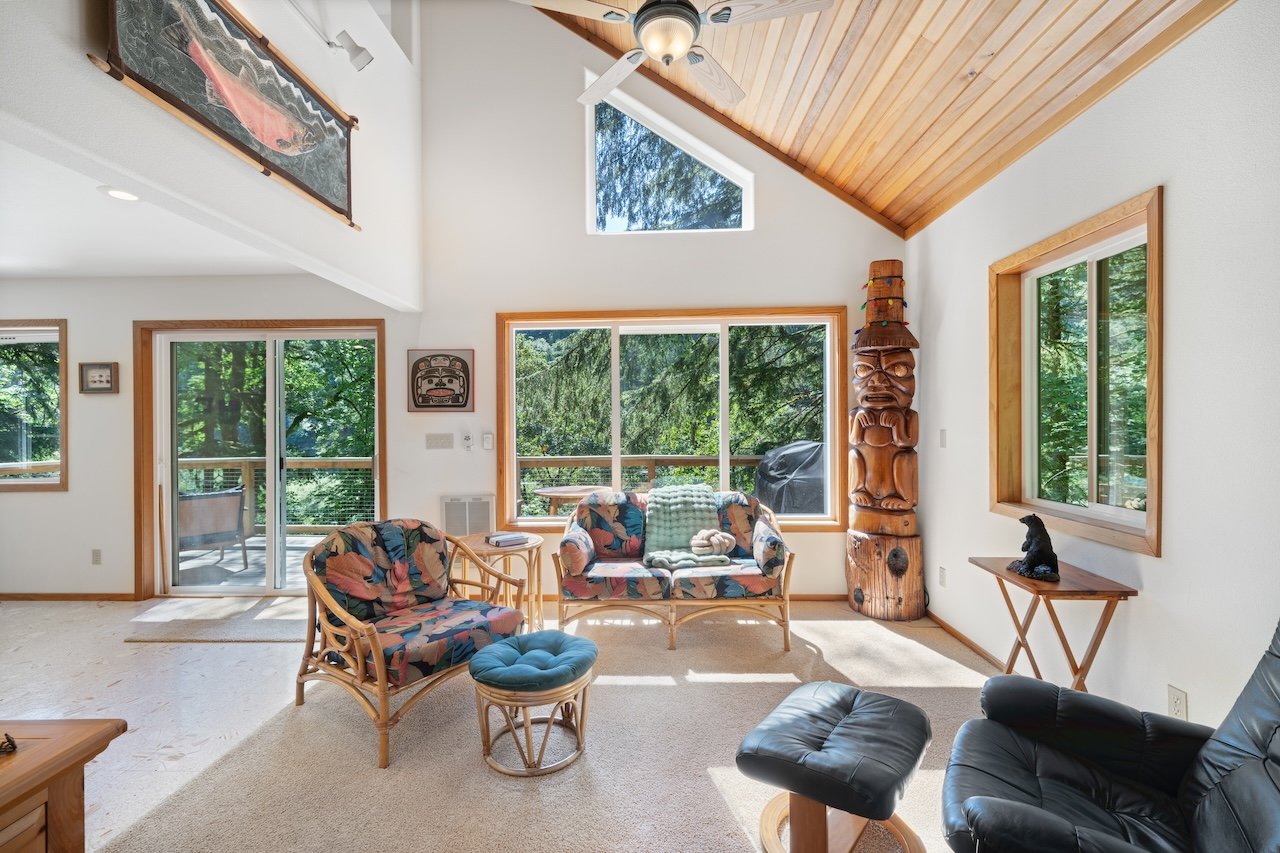 Bright living room with large windows, wooden ceiling, bamboo furniture, black leather chair, decorative totem pole, and outdoor view of trees.