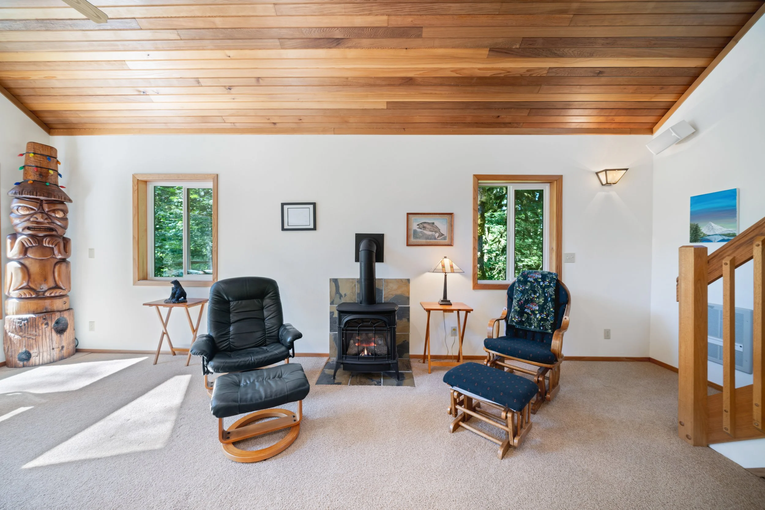 Living room with wood-paneled ceiling, fireplace, two windows showing green trees outside, black leather armchair, wooden side table, lamp, decorative art, and wooden staircase.