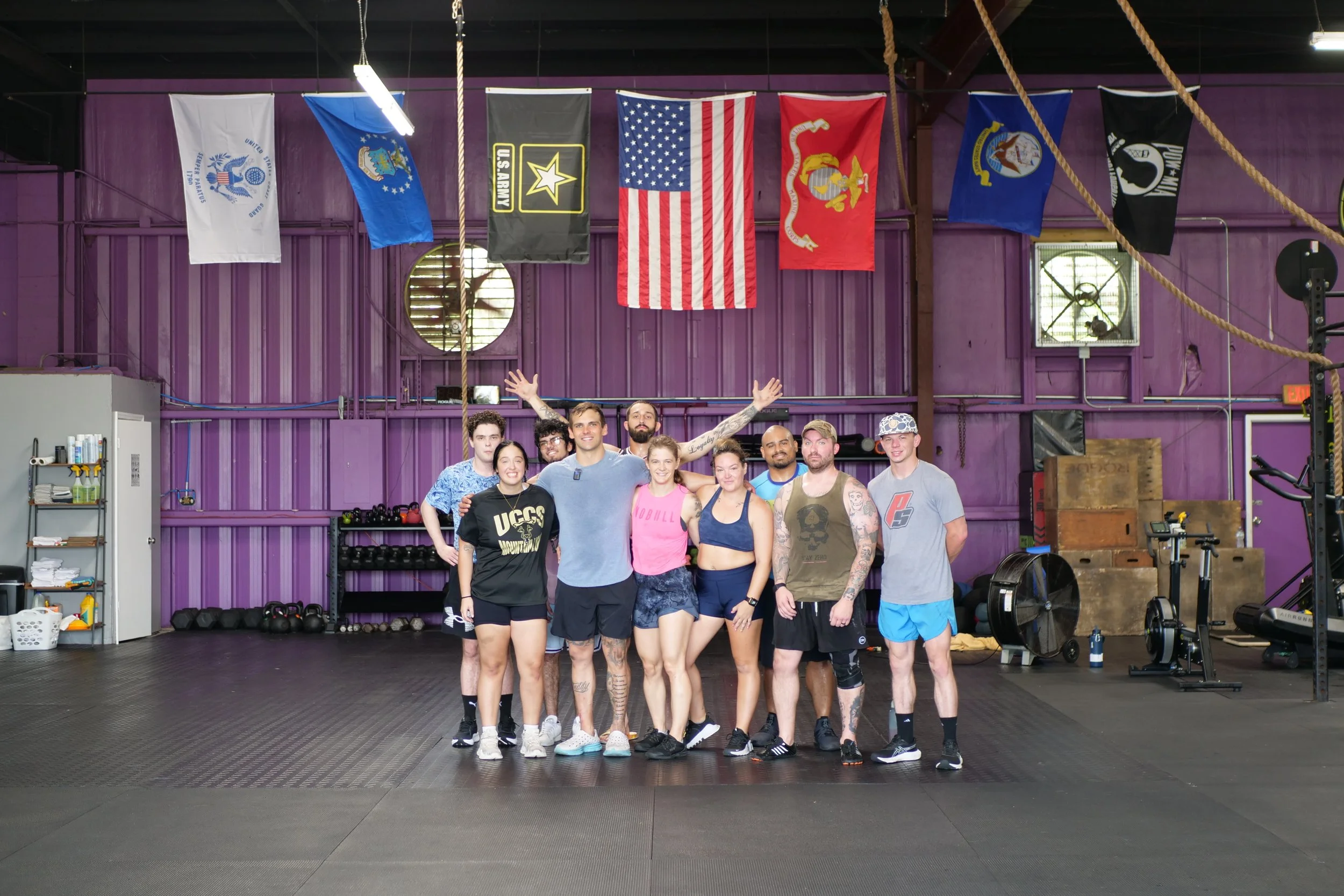 Group of nine people standing in a gym with purple walls and American flags hanging above. The group appears to be celebrating or posing after a workout, with some smiling and others making gestures. The gym has workout equipment and ropes visible in the background.