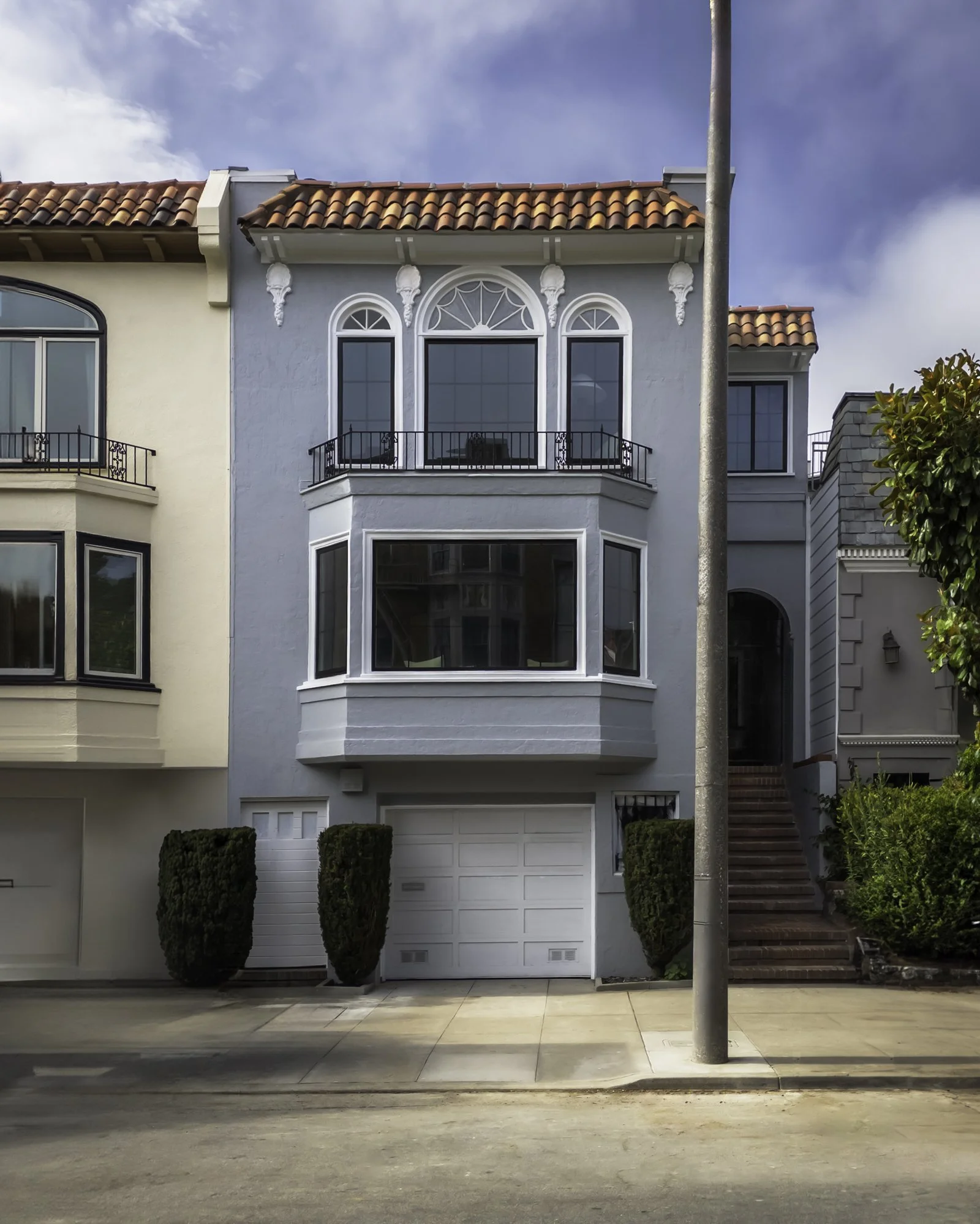 Front view of a two-story house with a white garage door, white and light blue exterior walls, black railing balcony on the second floor, and a red tile roof. There is a sidewalk in front with a street and a pole in the foreground.