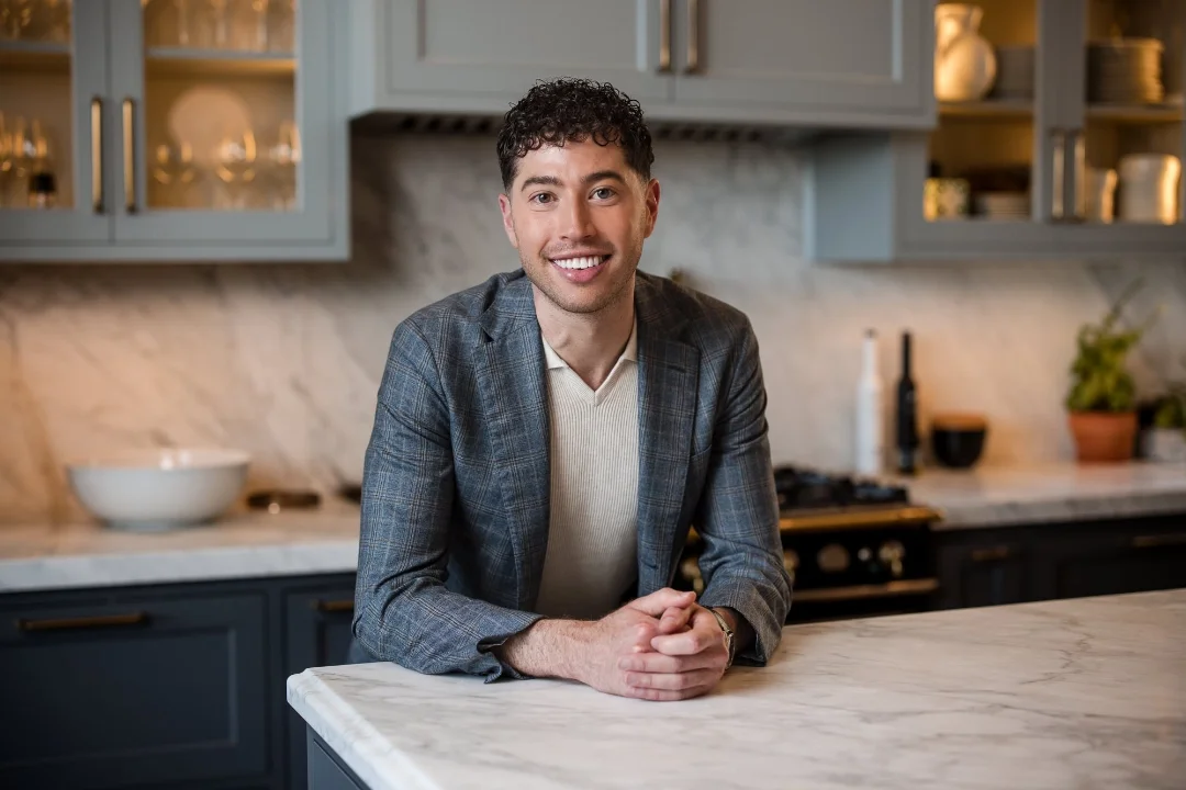 A young man with dark curly hair and a light complexion smiling and leaning on a white marble kitchen island.