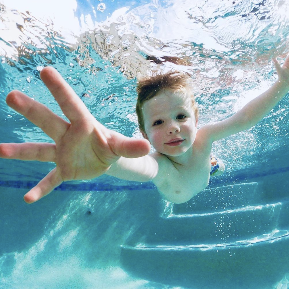 A young boy swimming underwater in a pool, reaching towards the camera with one hand.