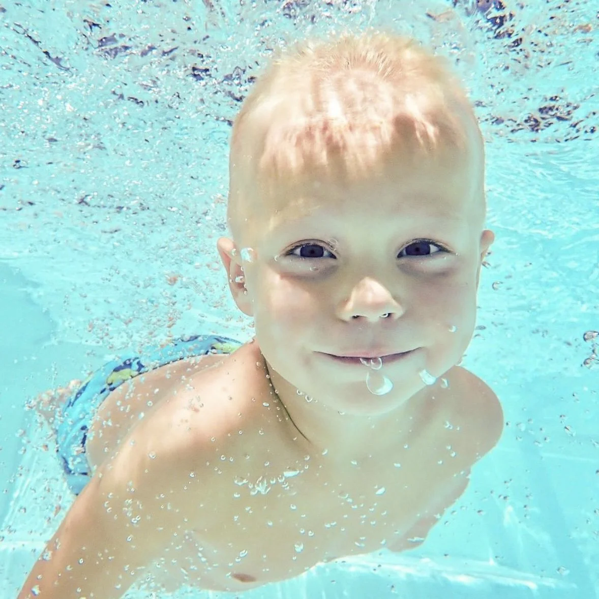 A young boy swimming underwater in a pool, smiling at the camera.
