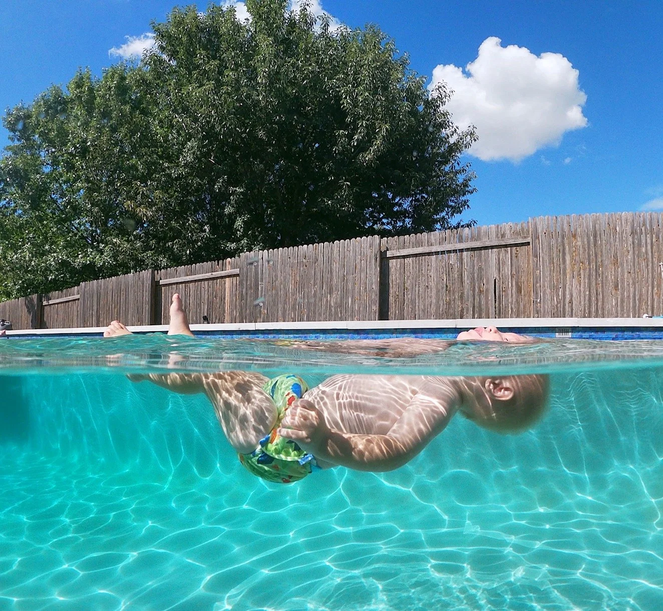 Person floating in swimming pool wearing colorful swim trunks on a sunny day with blue sky, trees, and wooden fence in the background.