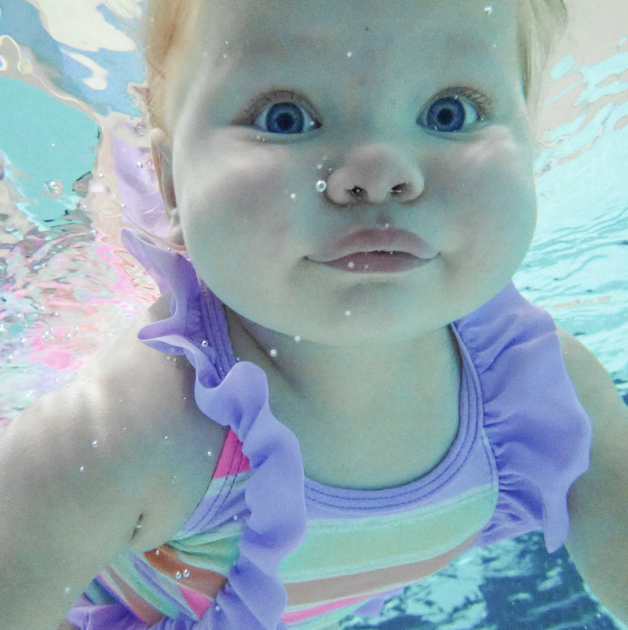 A young girl underwater in a swimming pool, wearing a colorful swimsuit with ruffles and smiling at the camera.
