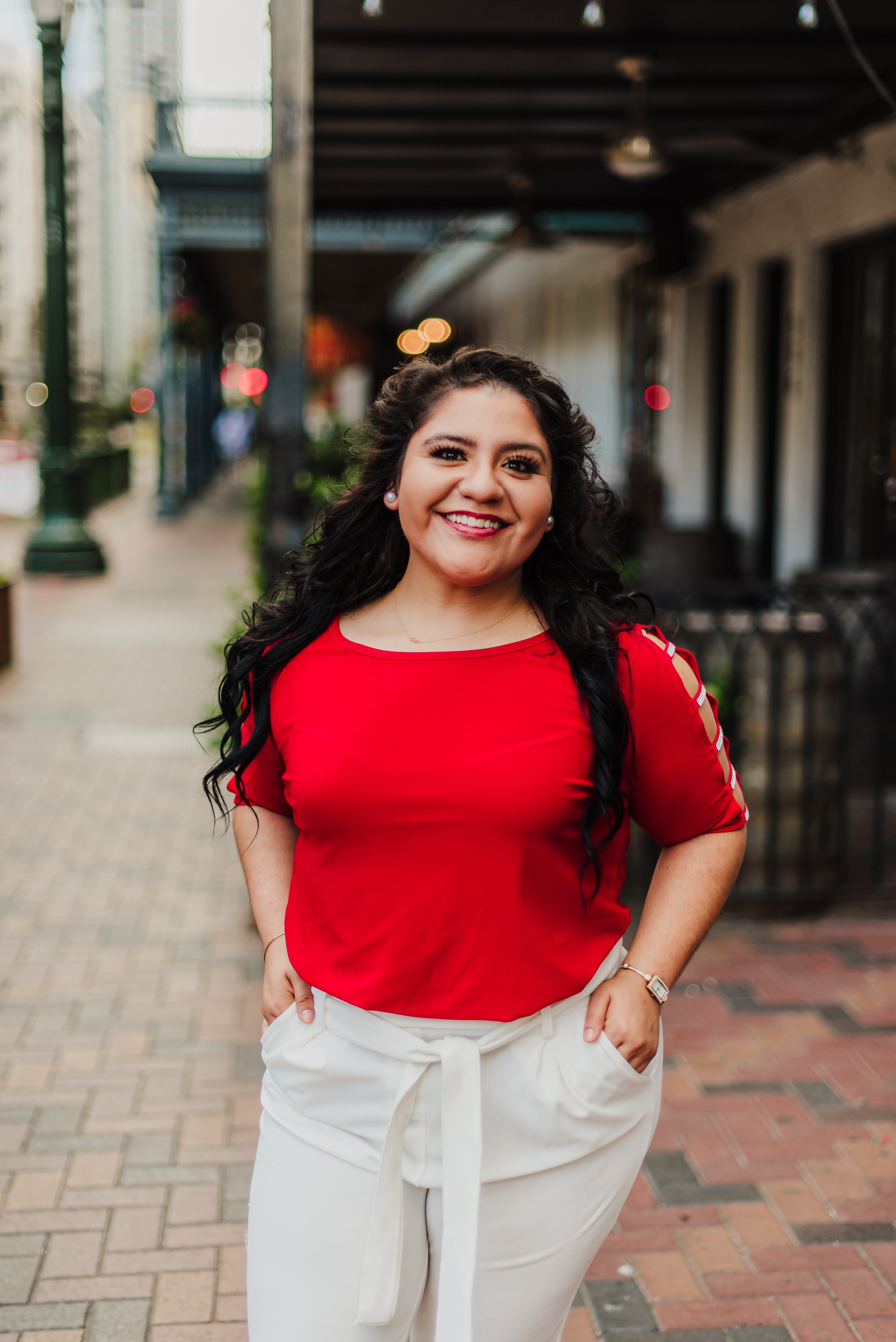 A woman with long dark curly hair, wearing a red top with cutout shoulders and white pants with a tied waist, standing on a city sidewalk, smiling at the camera.