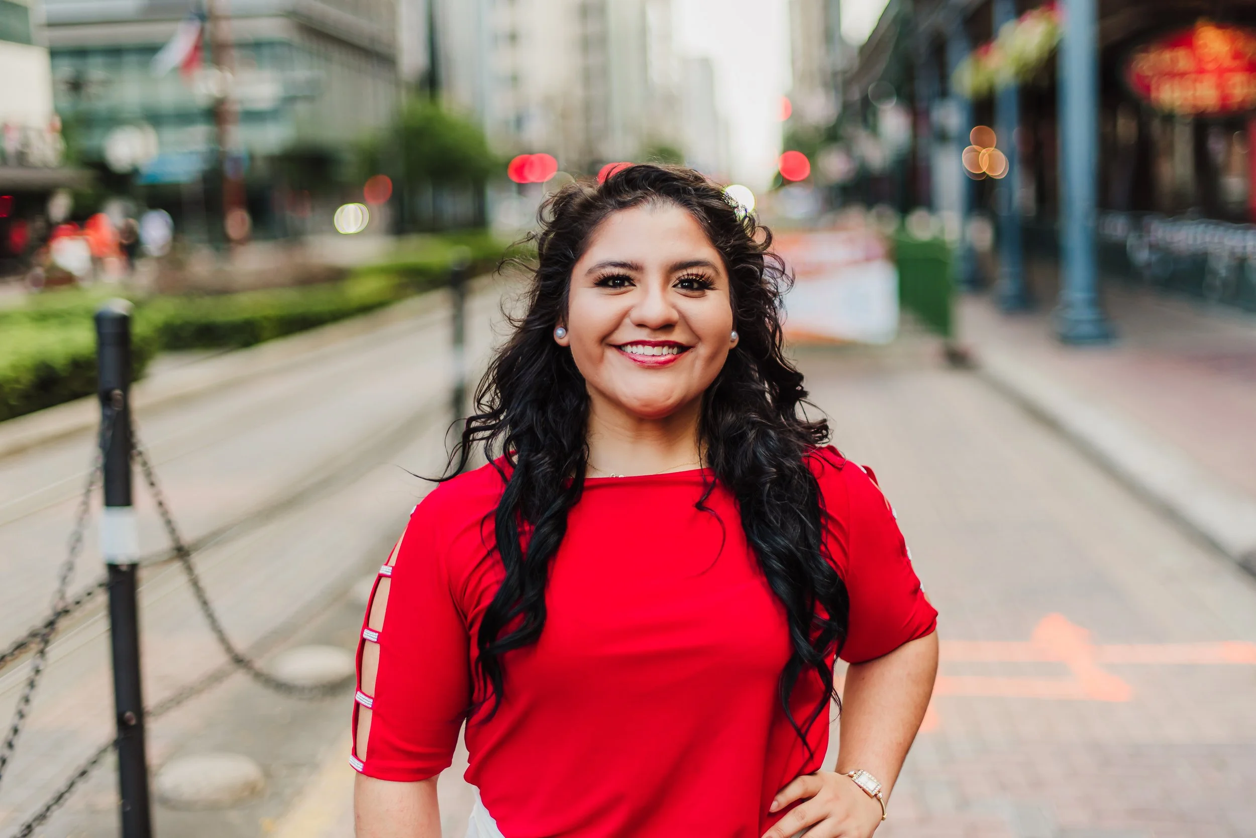 A woman in a red shirt smiling outdoors on a city street, with blurred buildings and colorful lights in the background.